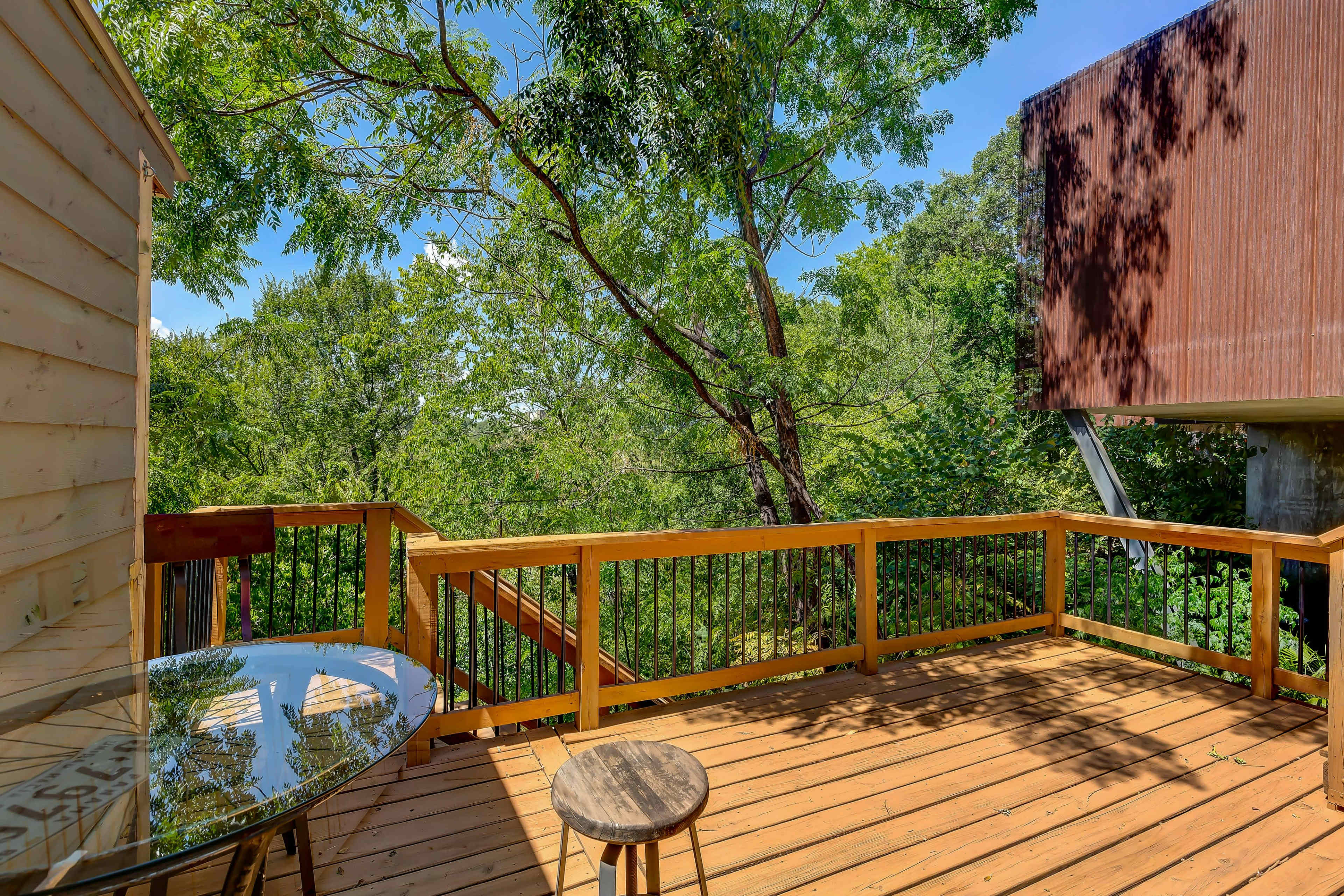 A wooden deck features a round table and stool, surrounded by greenery and trees.