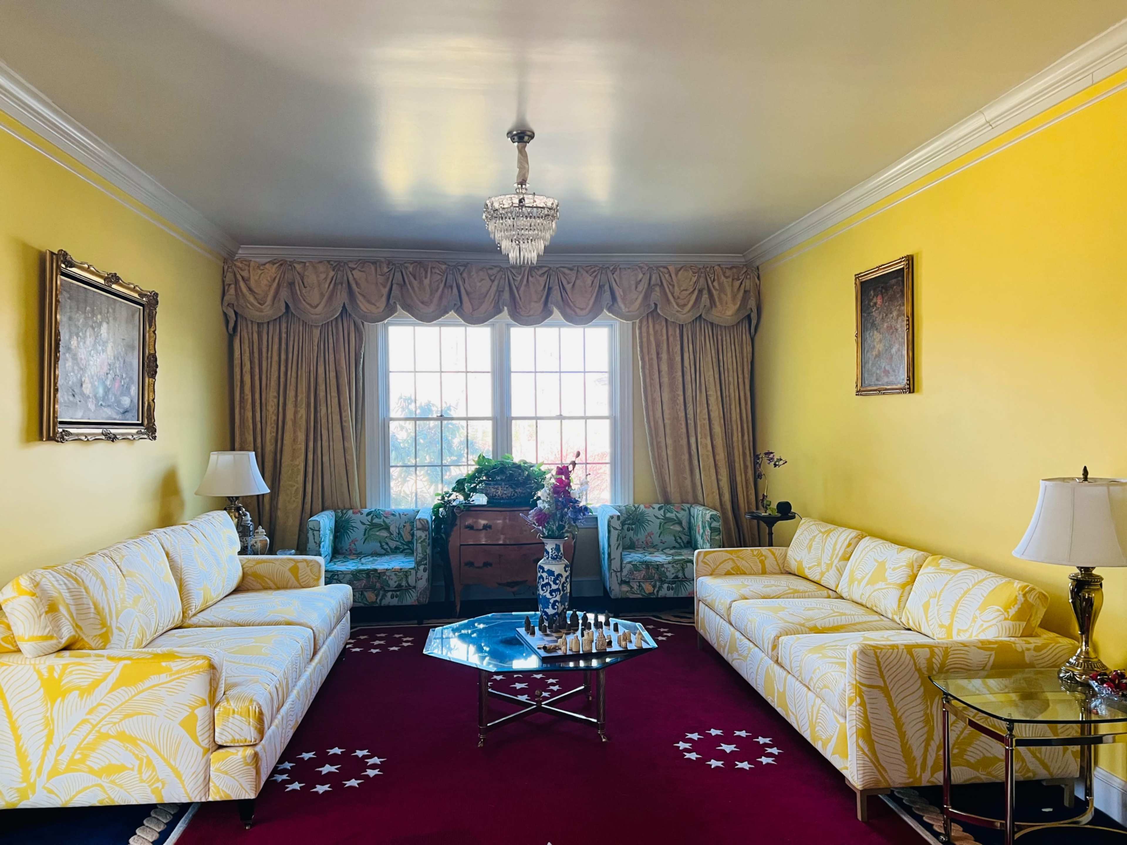 The living room features two yellow patterned sofas opposite each other, a glass coffee table in the center, and large windows draped with yellow curtains.
