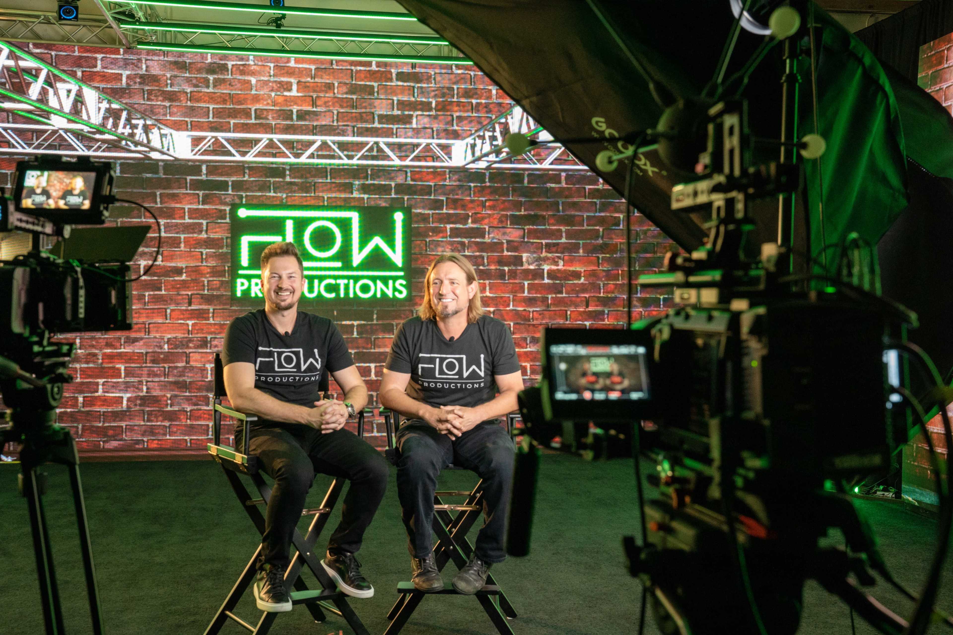Two men sit in director's chairs in front of a brick backdrop with the words "FLOW PRODUCTIONS" lit up in green.