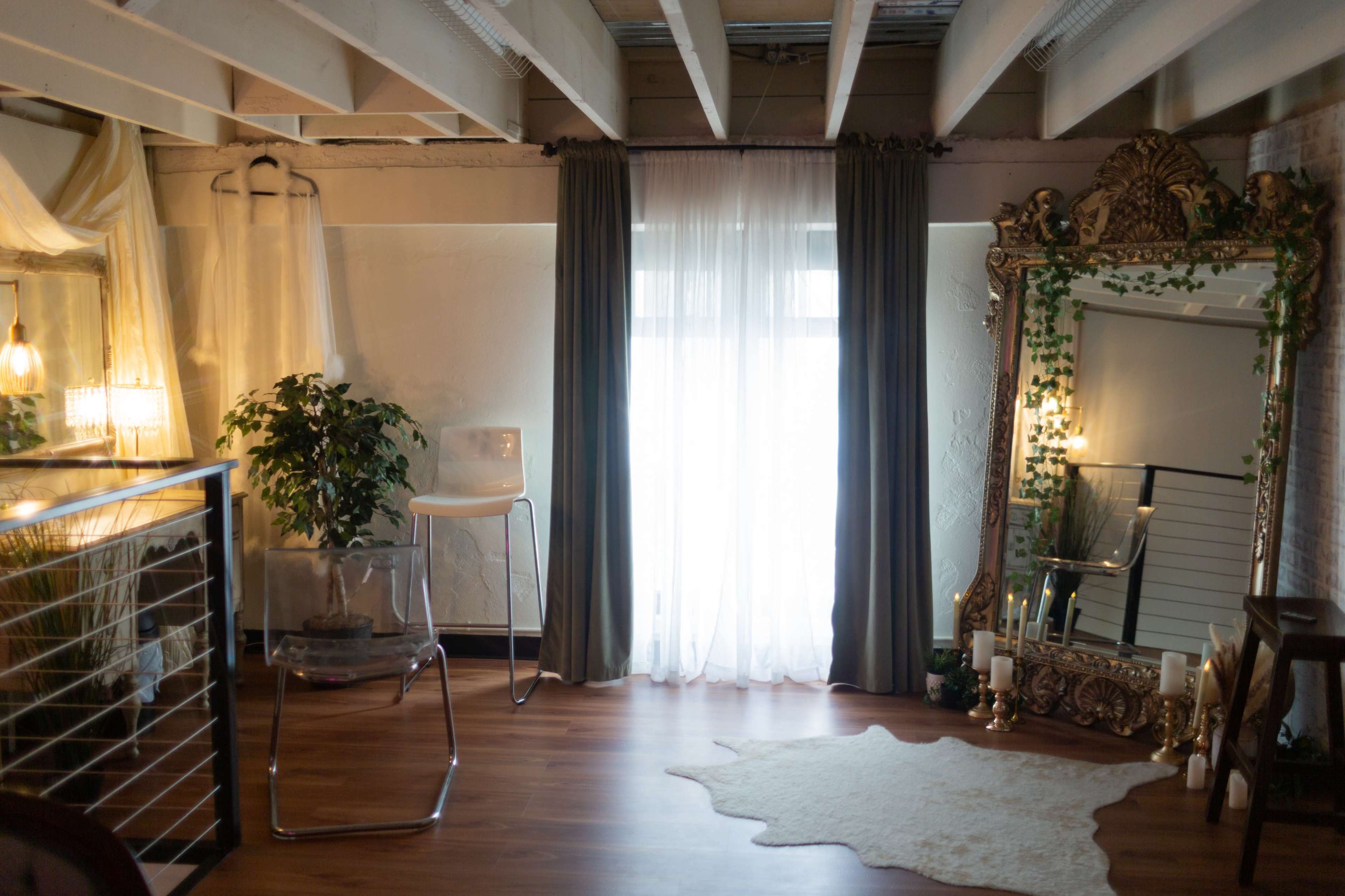 The scene features a stylish interior with large windows adorned by sheer curtains, a large decorative mirror, and a white area rug on the wooden floor.
