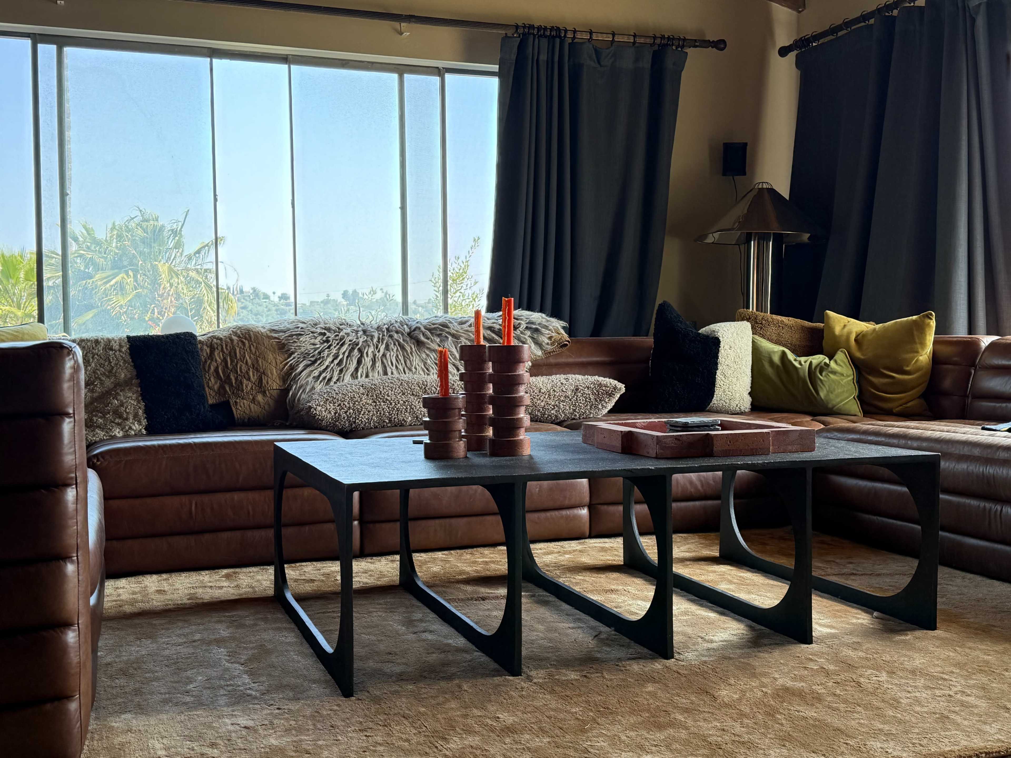 The scene shows a modern living room featuring a brown leather sectional sofa, a set of decorative candles on a black coffee table, and large windows allowing natural light to fill the space.