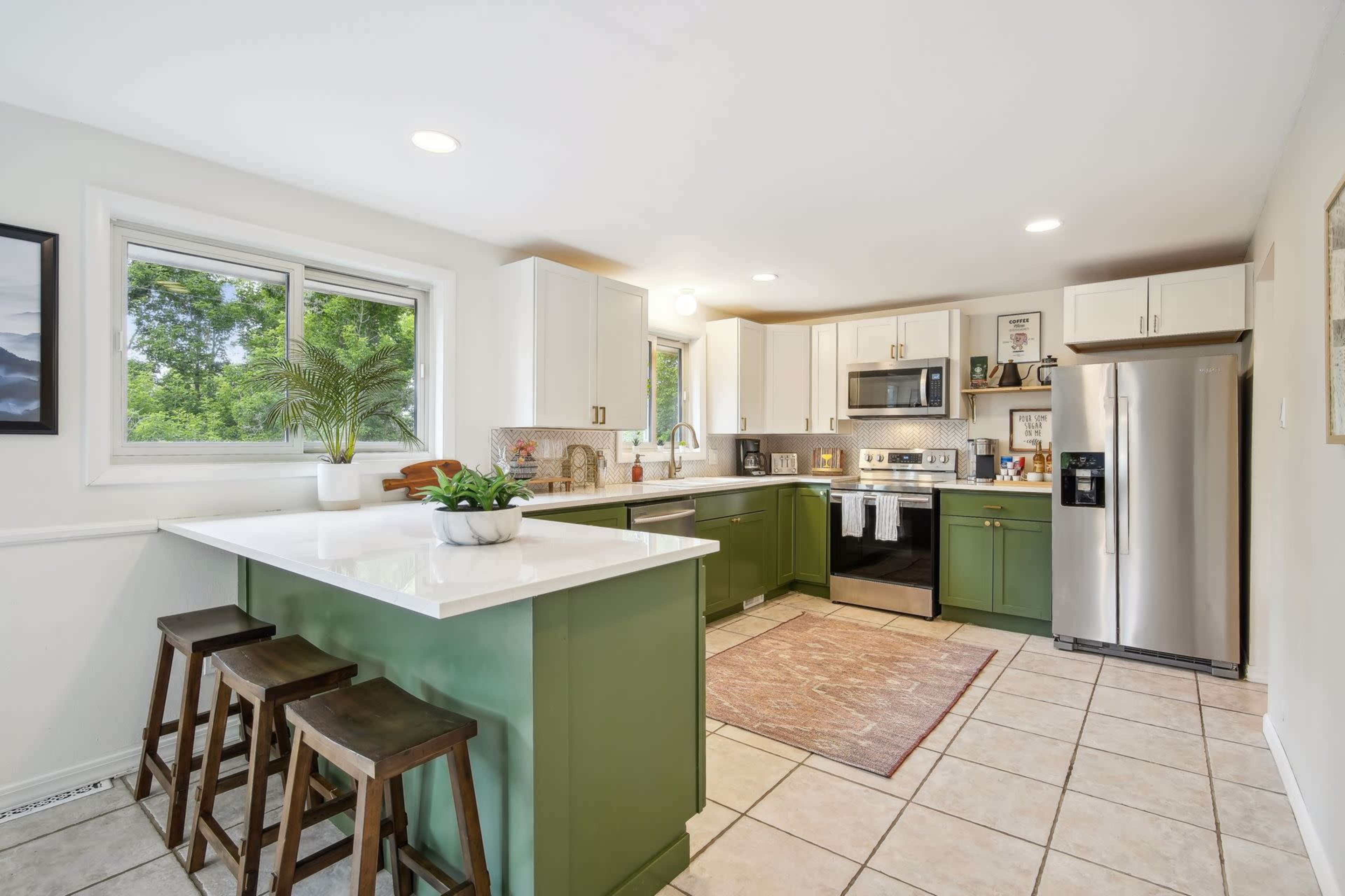 A bright kitchen features green cabinets, stainless steel appliances, and a large window overlooking greenery.