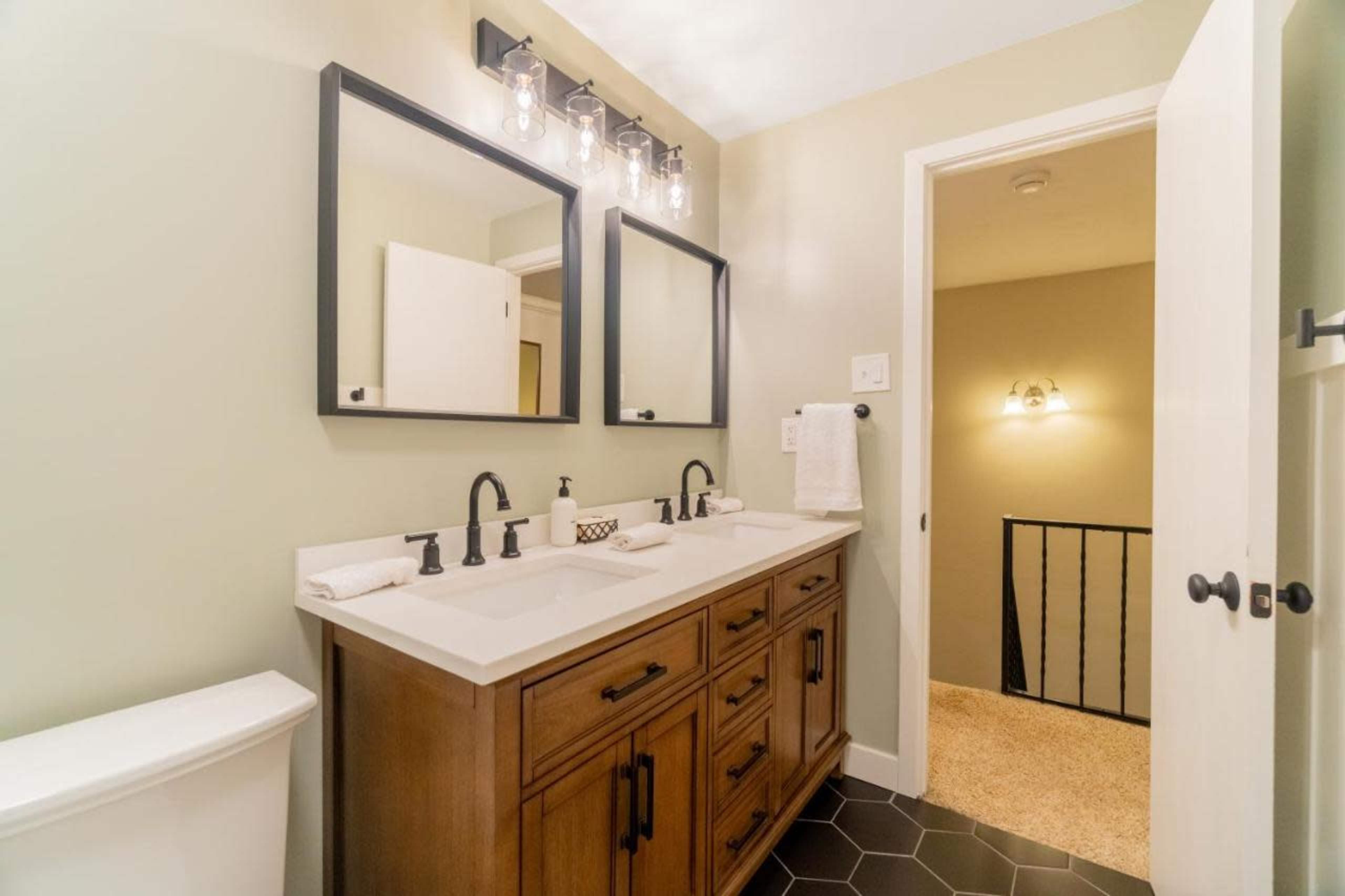 The image shows a bathroom featuring a double vanity with two sinks, modern light fixtures, and two mirrors above the sink area, with a door leading to a staircase.