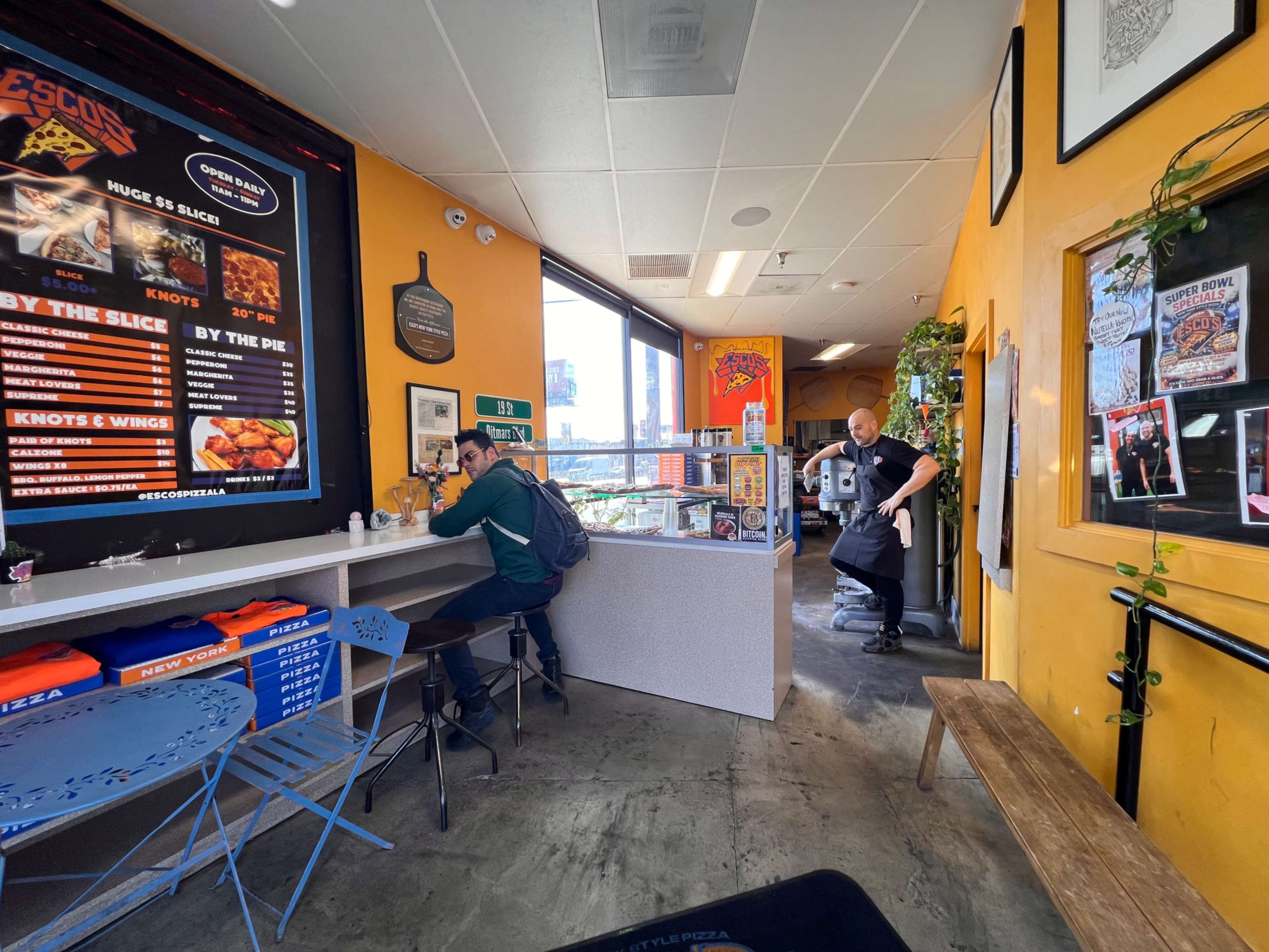 The interior of a restaurant features a colorful decor, a person seated at a counter, and an employee preparing food in the background.