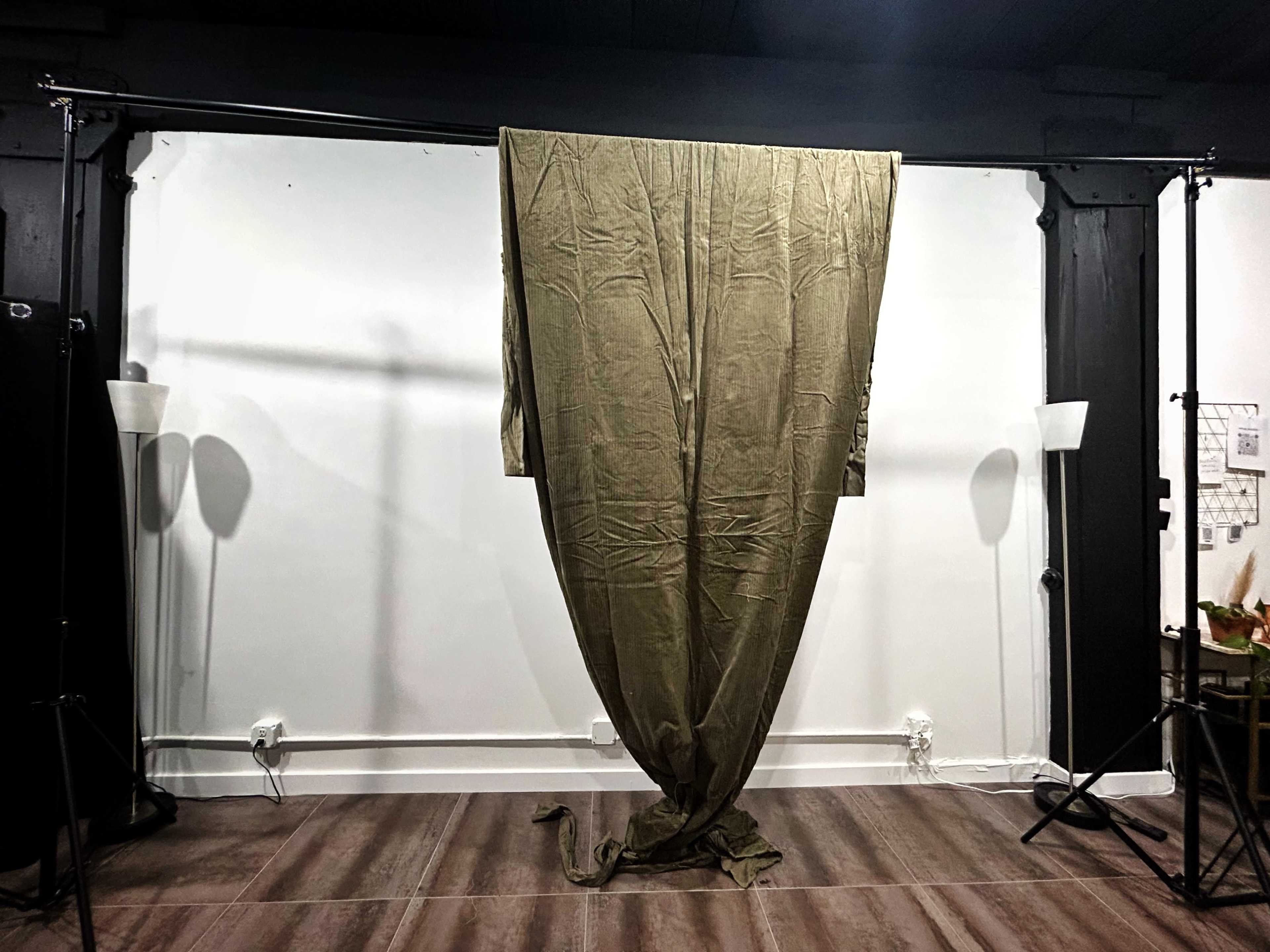 The image shows a large, olive-green fabric hanging from a support structure against a plain white wall, with two lamps positioned on the sides.