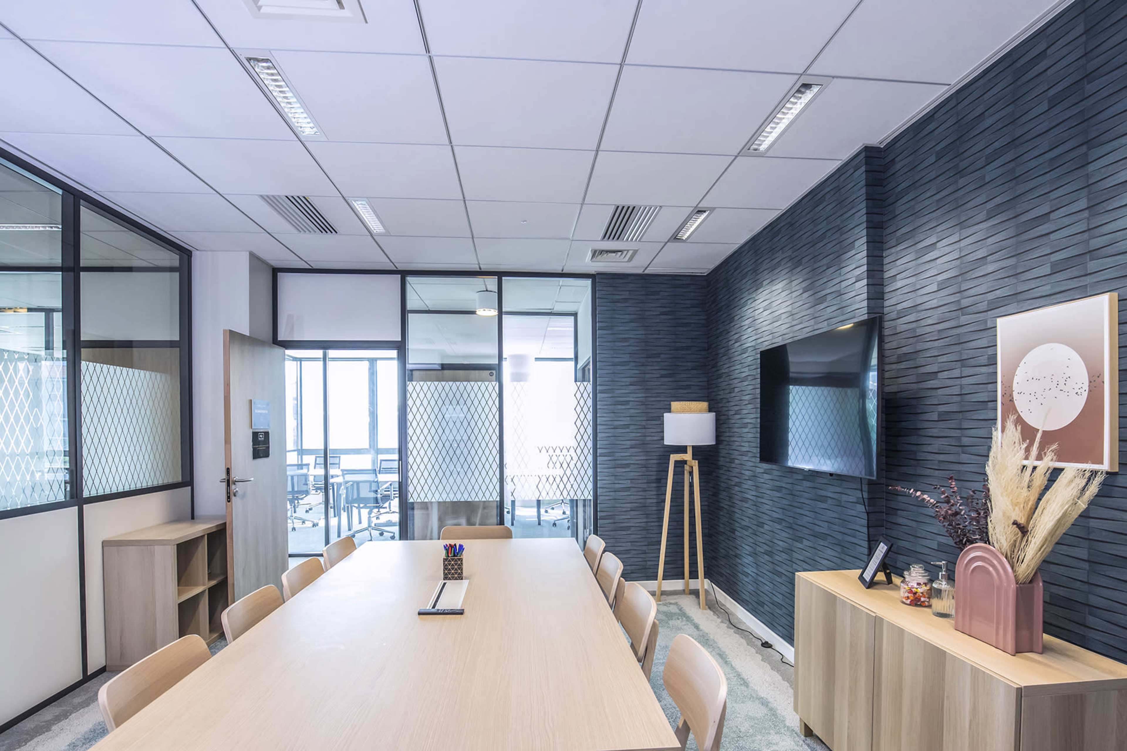 A modern meeting room features a long wooden table surrounded by chairs, glass walls, and a wall-mounted TV.