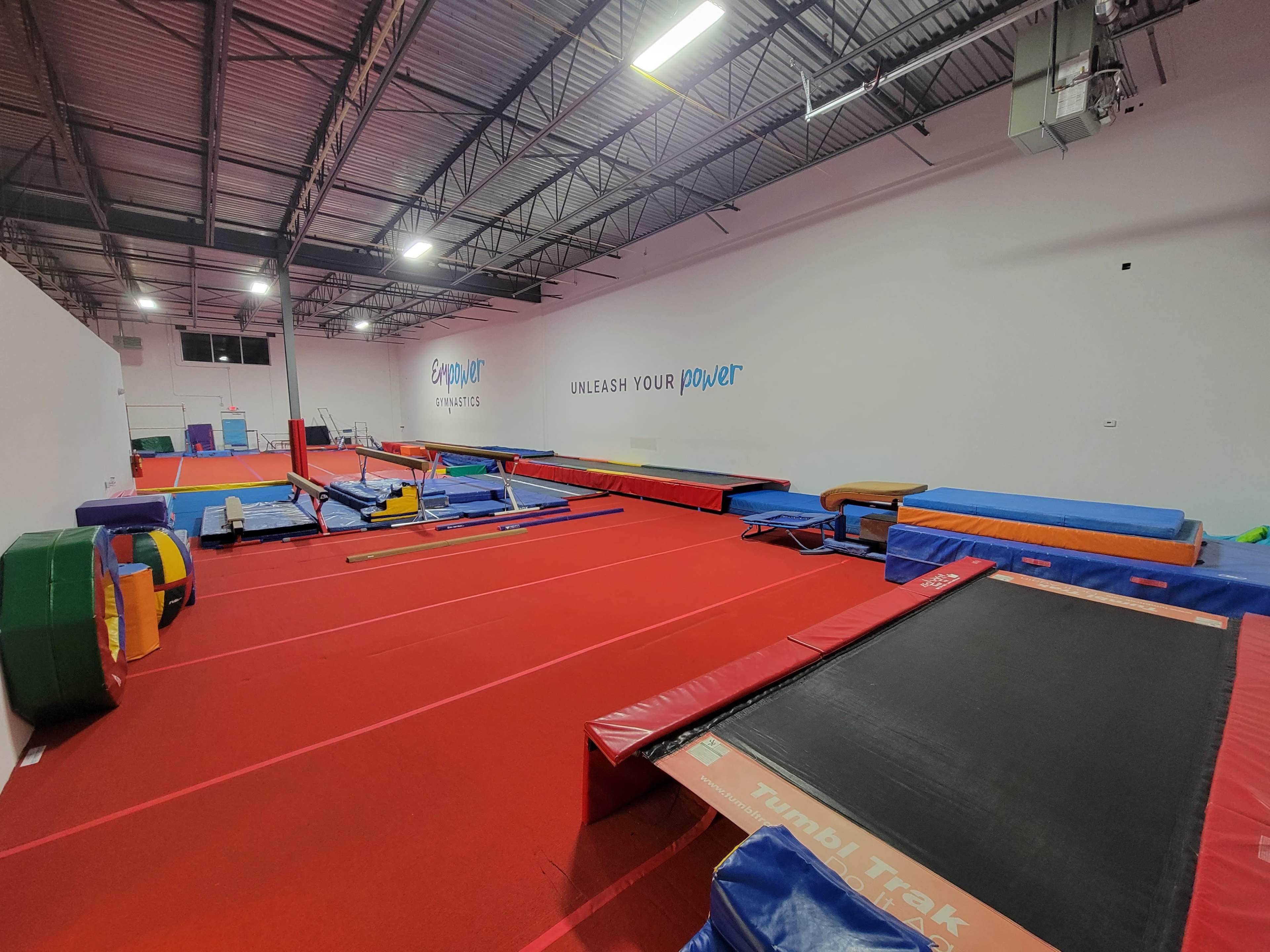 A spacious gymnastics training facility equipped with various mats, trampolines, and apparatuses on a red floor.