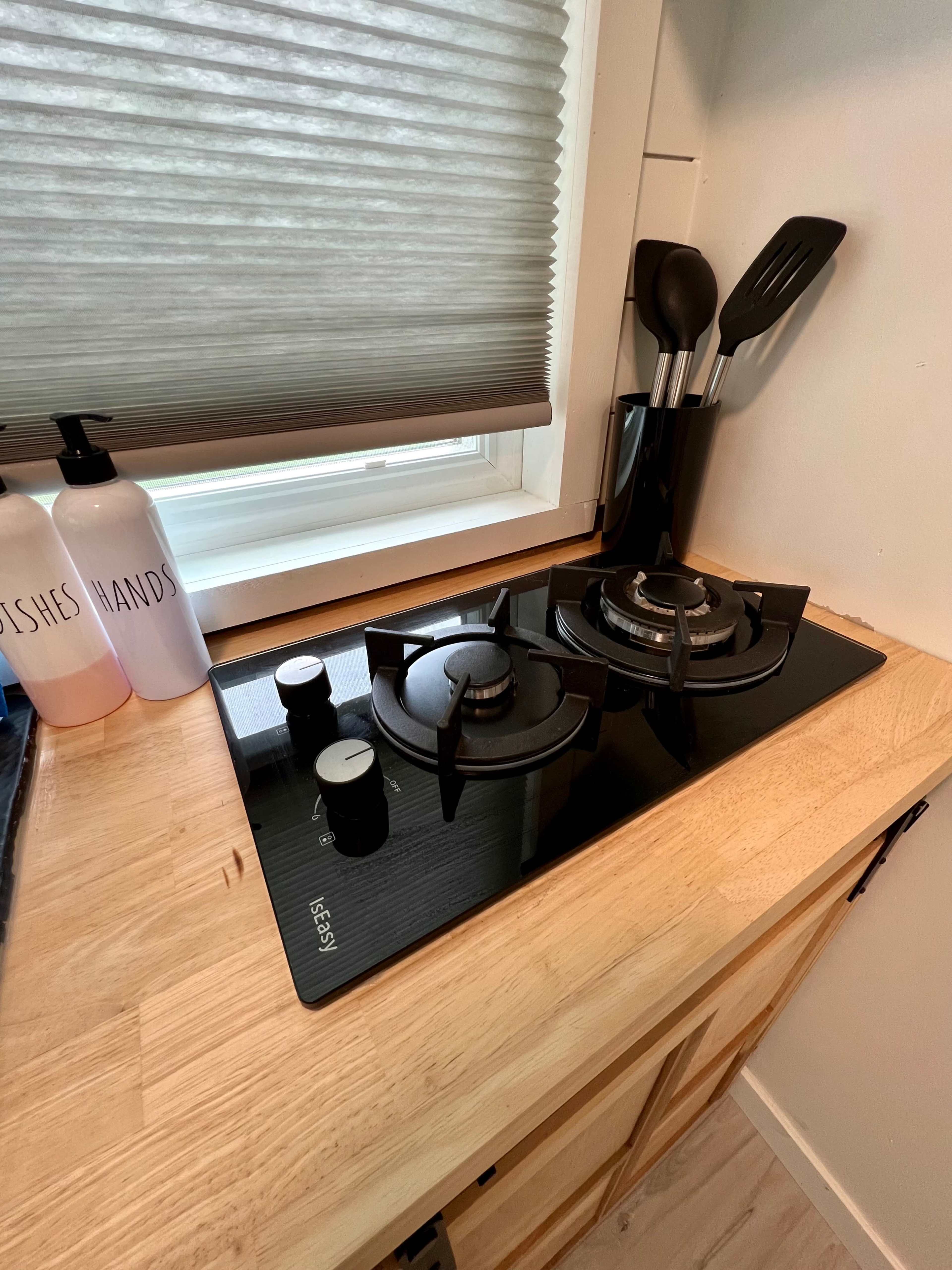 A black gas stovetop with two burners is installed on a wooden countertop near a window, alongside kitchen utensils in a holder and bottles labeled "DISHES" and "HANDS."
