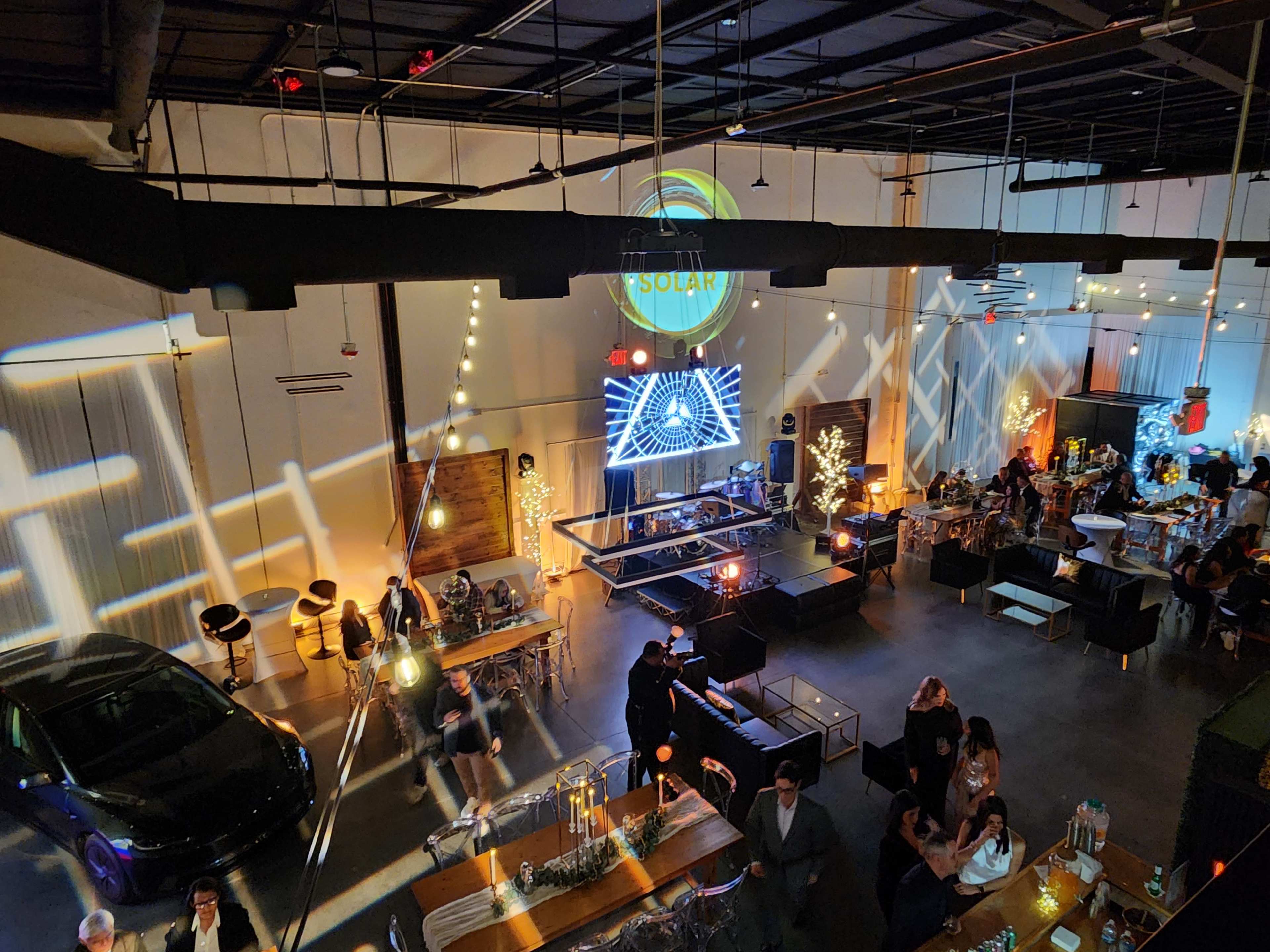 The scene shows a spacious event venue with tables and chairs, decorative lighting, and a stage featuring a large projection, with people mingling and a car displayed prominently in the background.