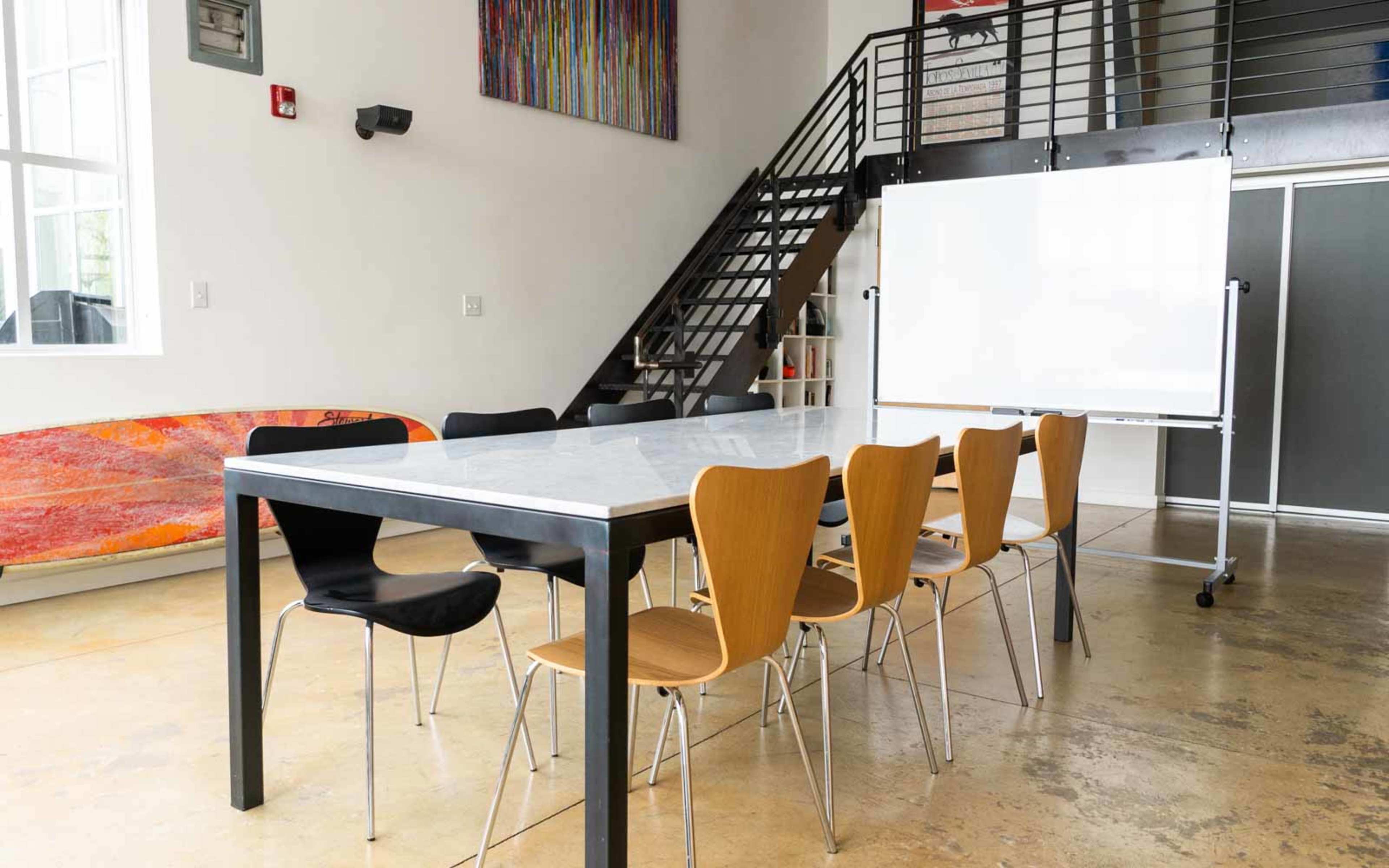 A modern meeting room with a long table and black and wood chairs, featuring a whiteboard on a stand and a staircase in the background.
