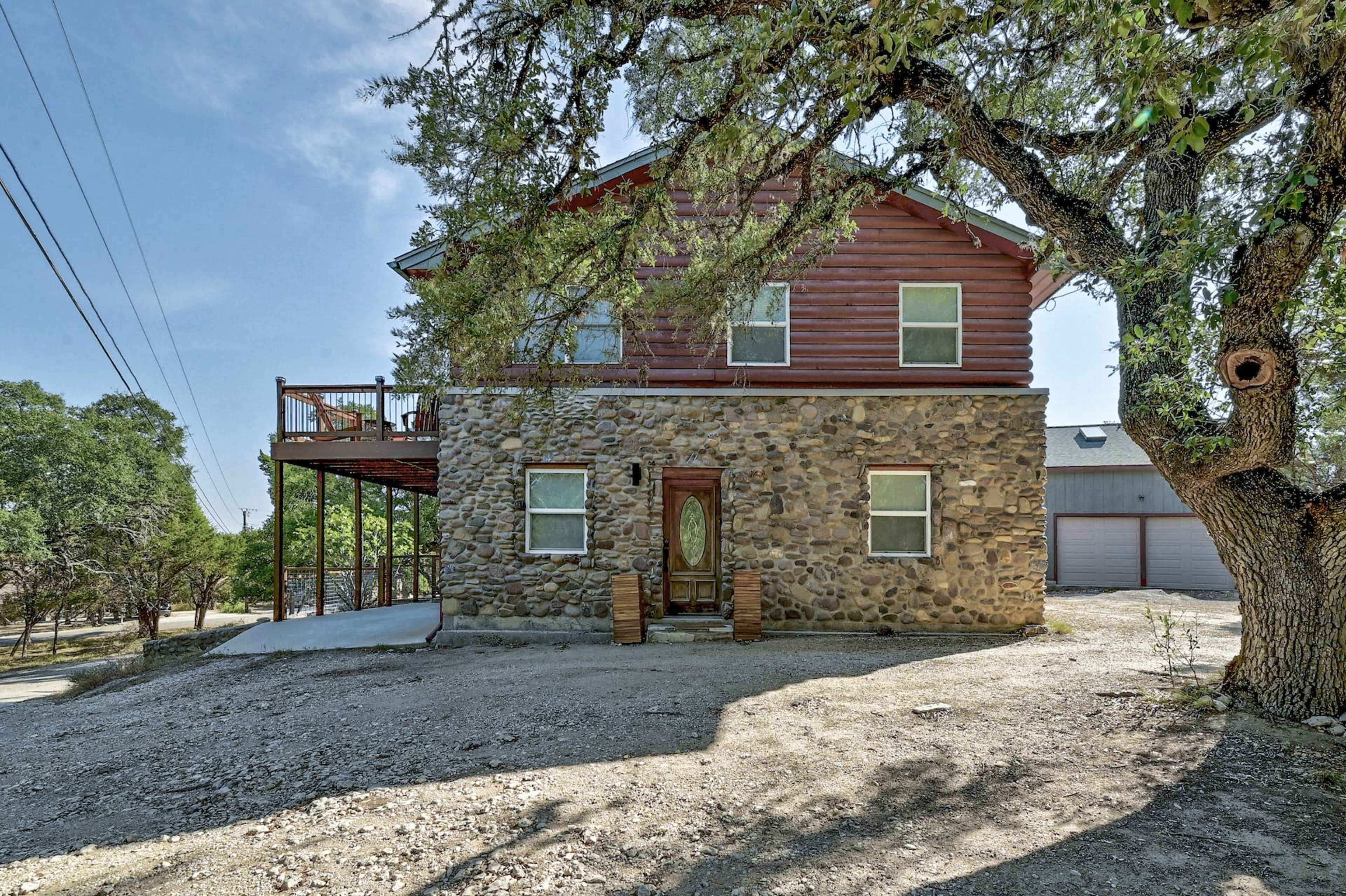 A three-story house with a stone-covered lower level and wooden upper section is situated under a tree, with a driveway and a garage visible in the background.