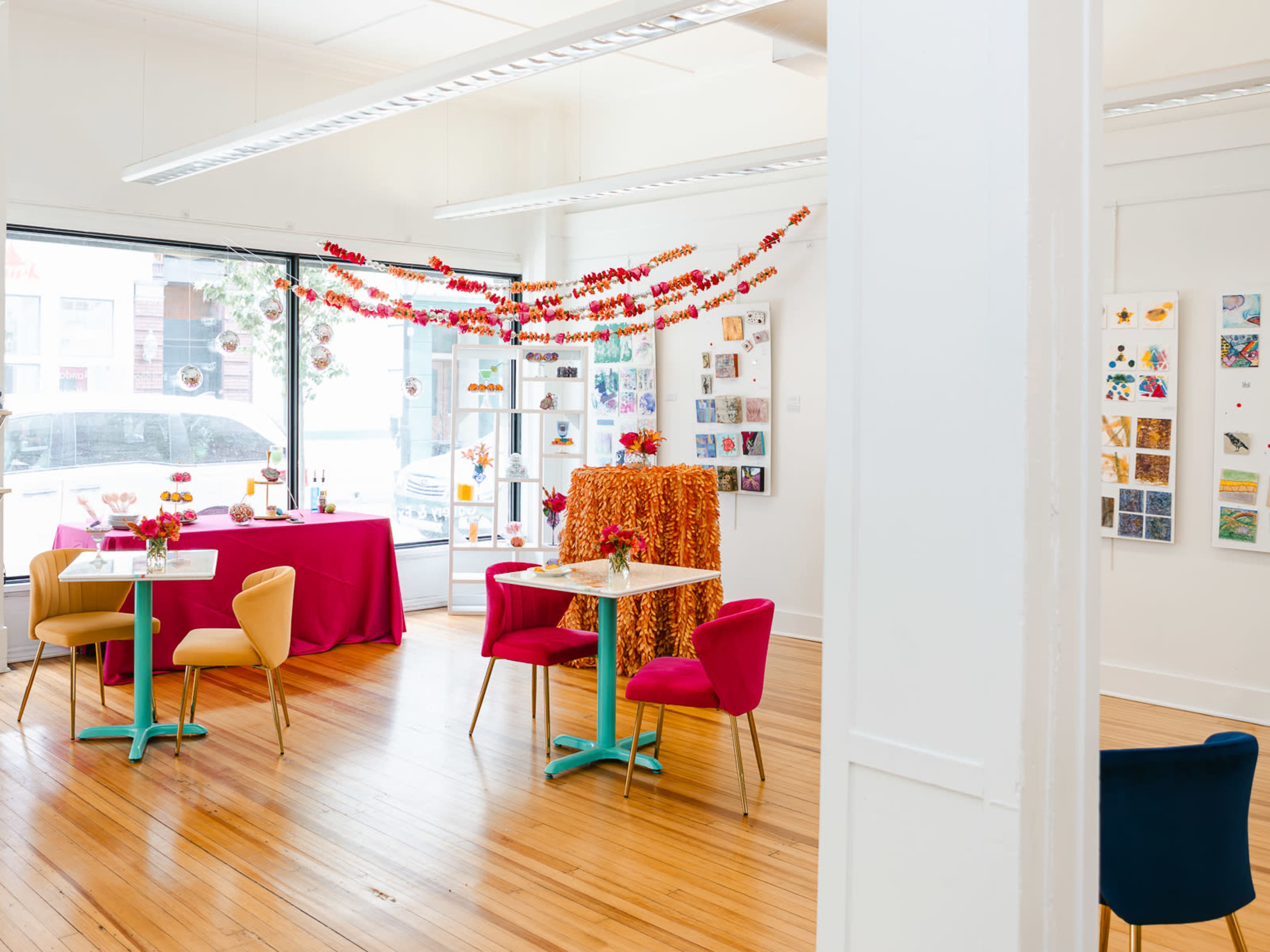 A brightly lit art gallery featuring colorful decorations, with tables and chairs arranged around a central area filled with displayed artwork on the walls.