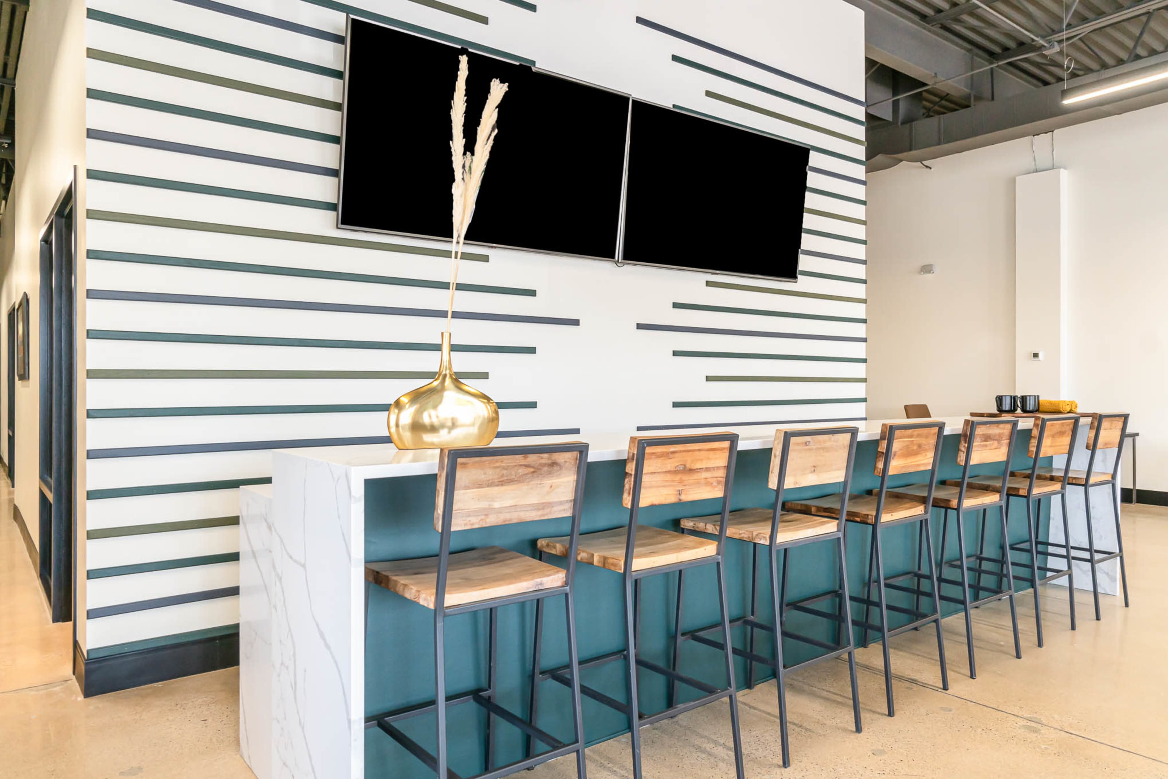A modern bar area with high wooden stools lined up against a striped wall, complemented by two blank television screens and a decorative vase.