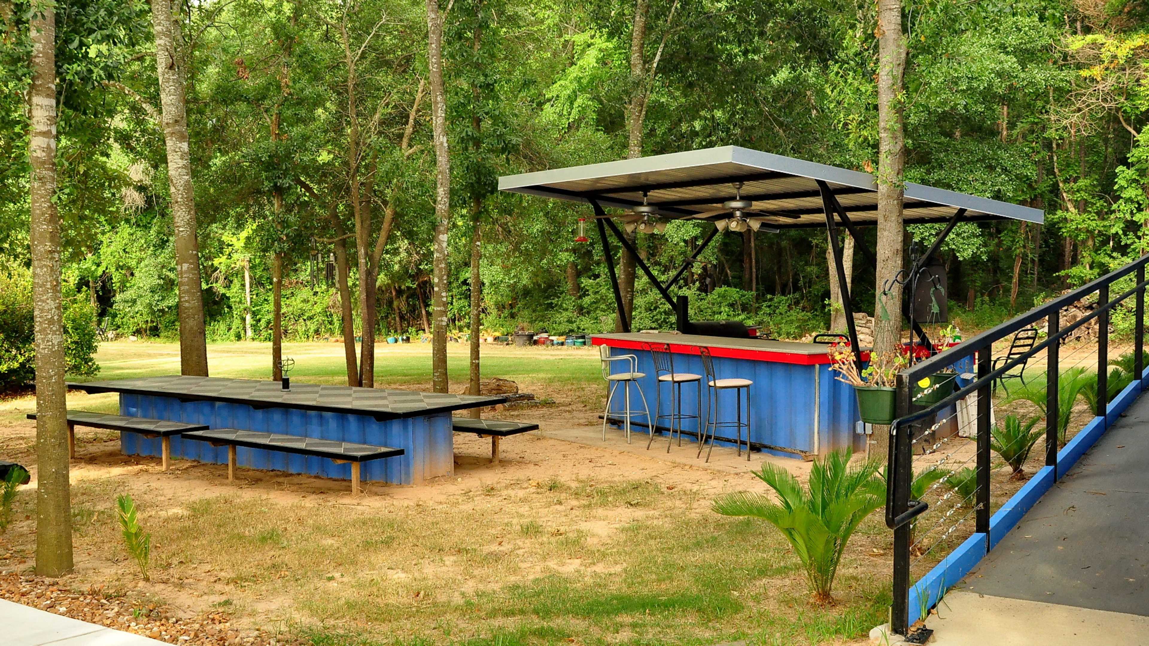 A blue outdoor bar with stools is set near a shaded picnic area surrounded by trees.