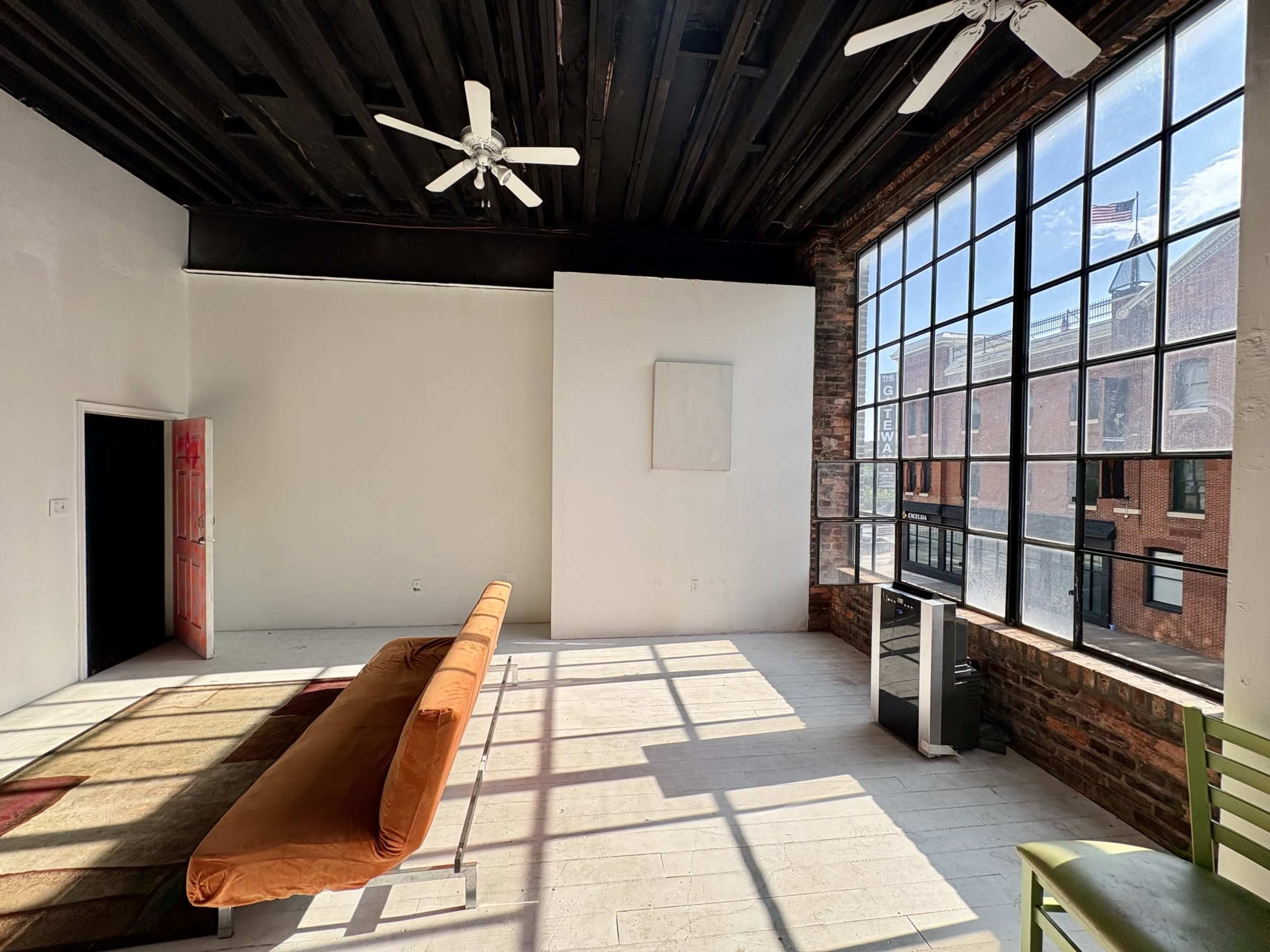 The image shows a bright, spacious room with large windows, a brown couch in the center, and a white wall with a door leading to another space.