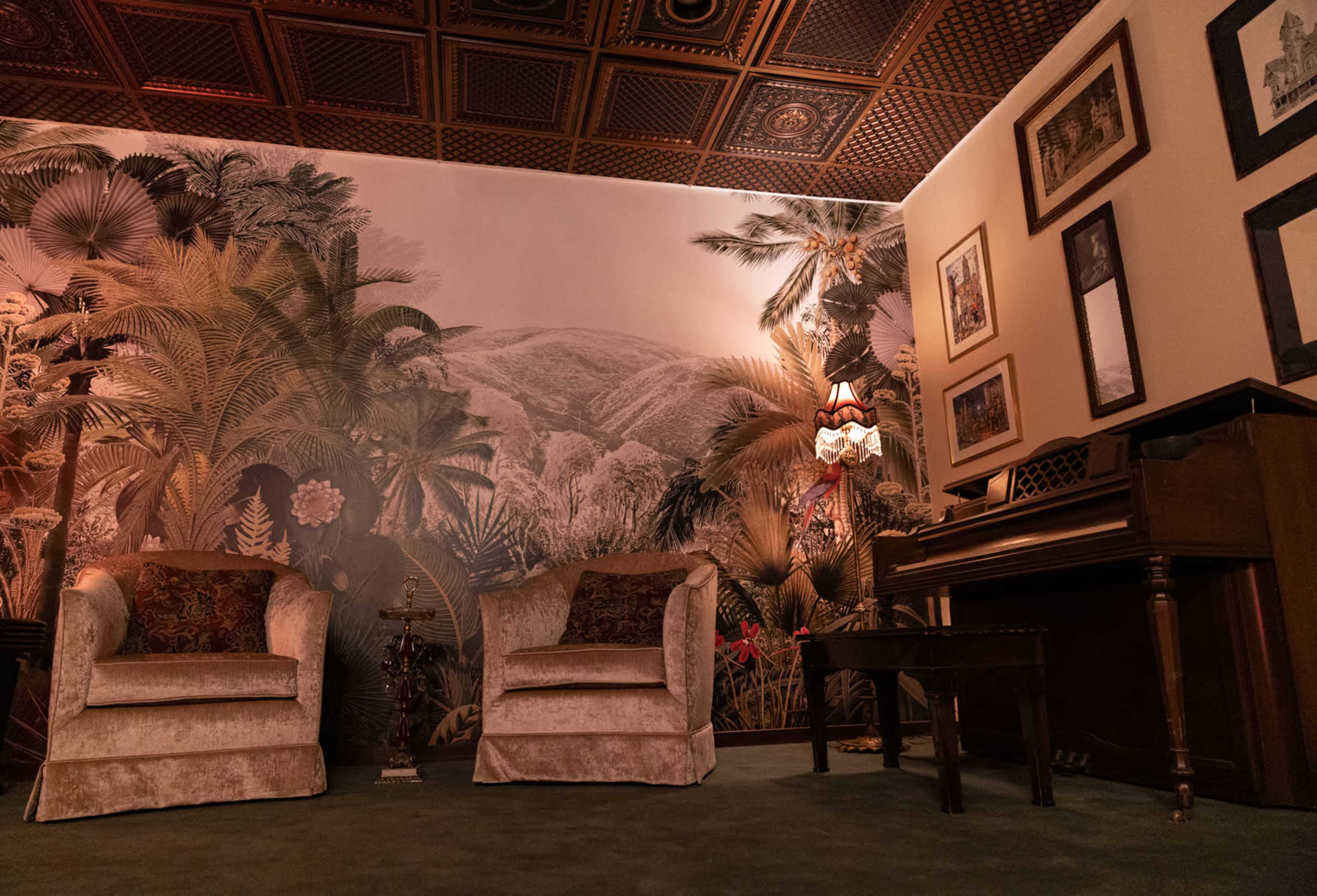 The image shows a cozy room decorated with a large wallpaper mural of tropical plants, featuring two plush armchairs and a vintage piano.