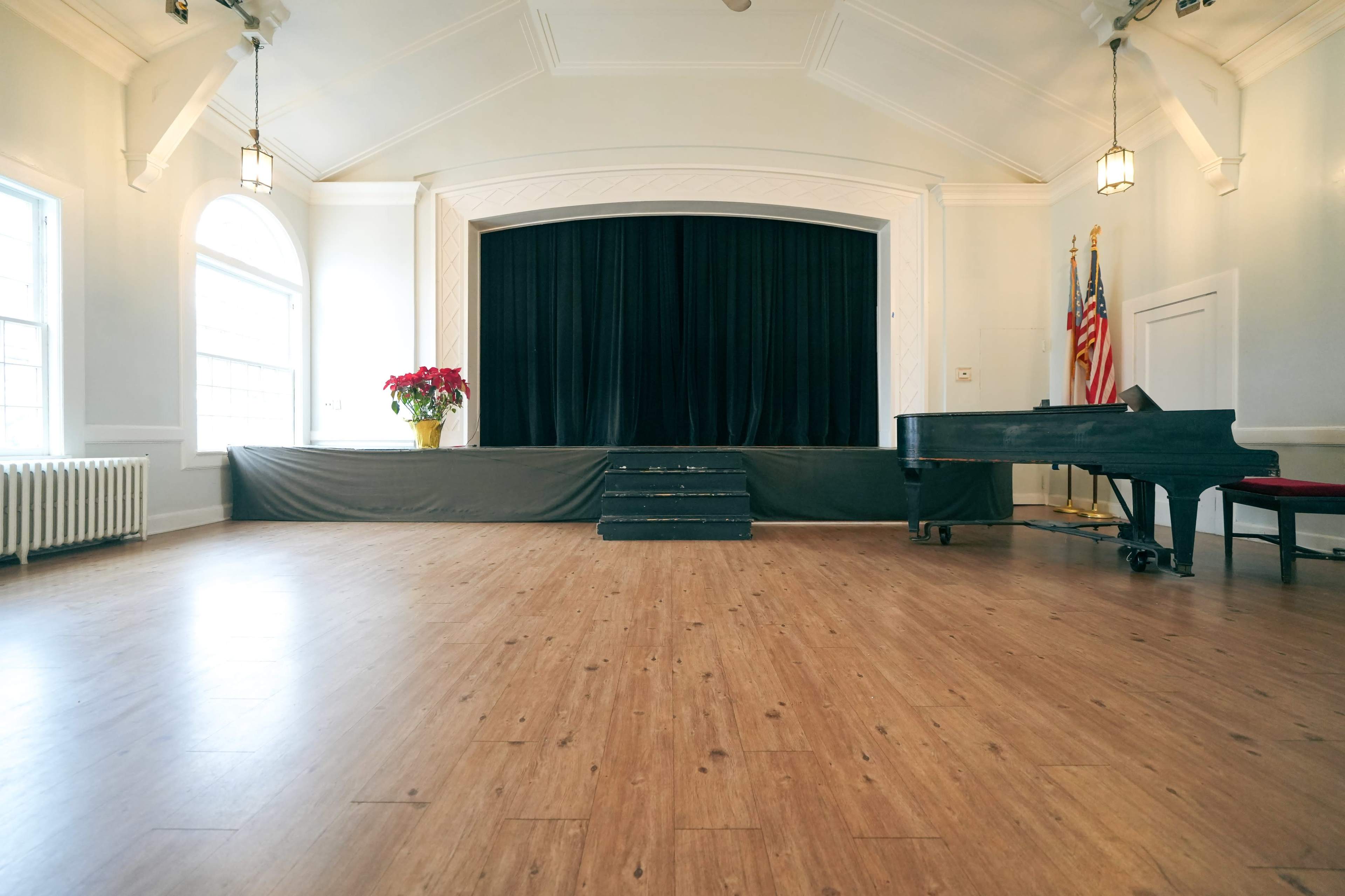 The image shows an empty performance stage with a black curtain, steps leading up to it, a grand piano to the right, and American flags in the background.