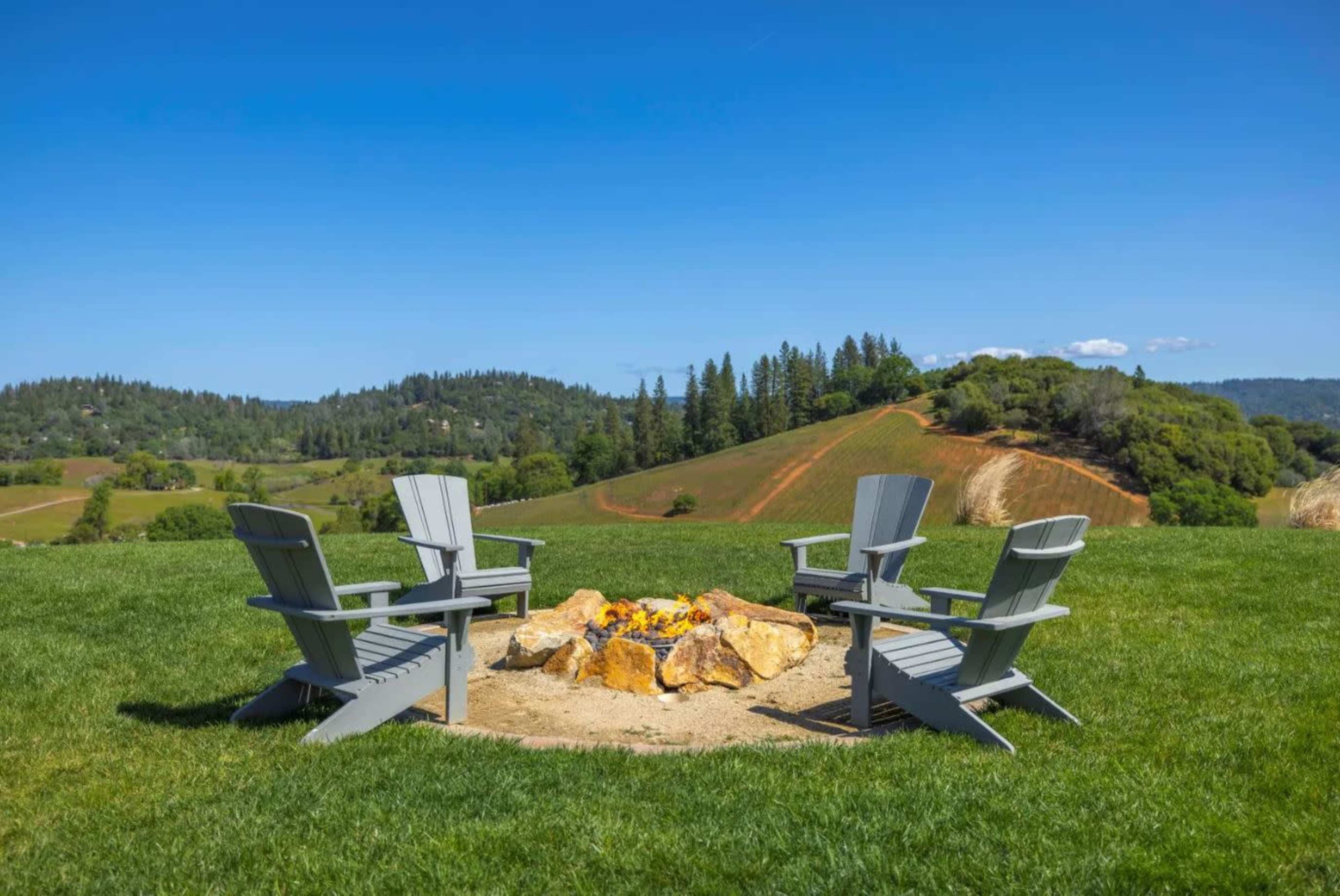 A circle of gray Adirondack chairs surrounds a fire pit made of large stones on a grassy area overlooking rolling hills and trees.