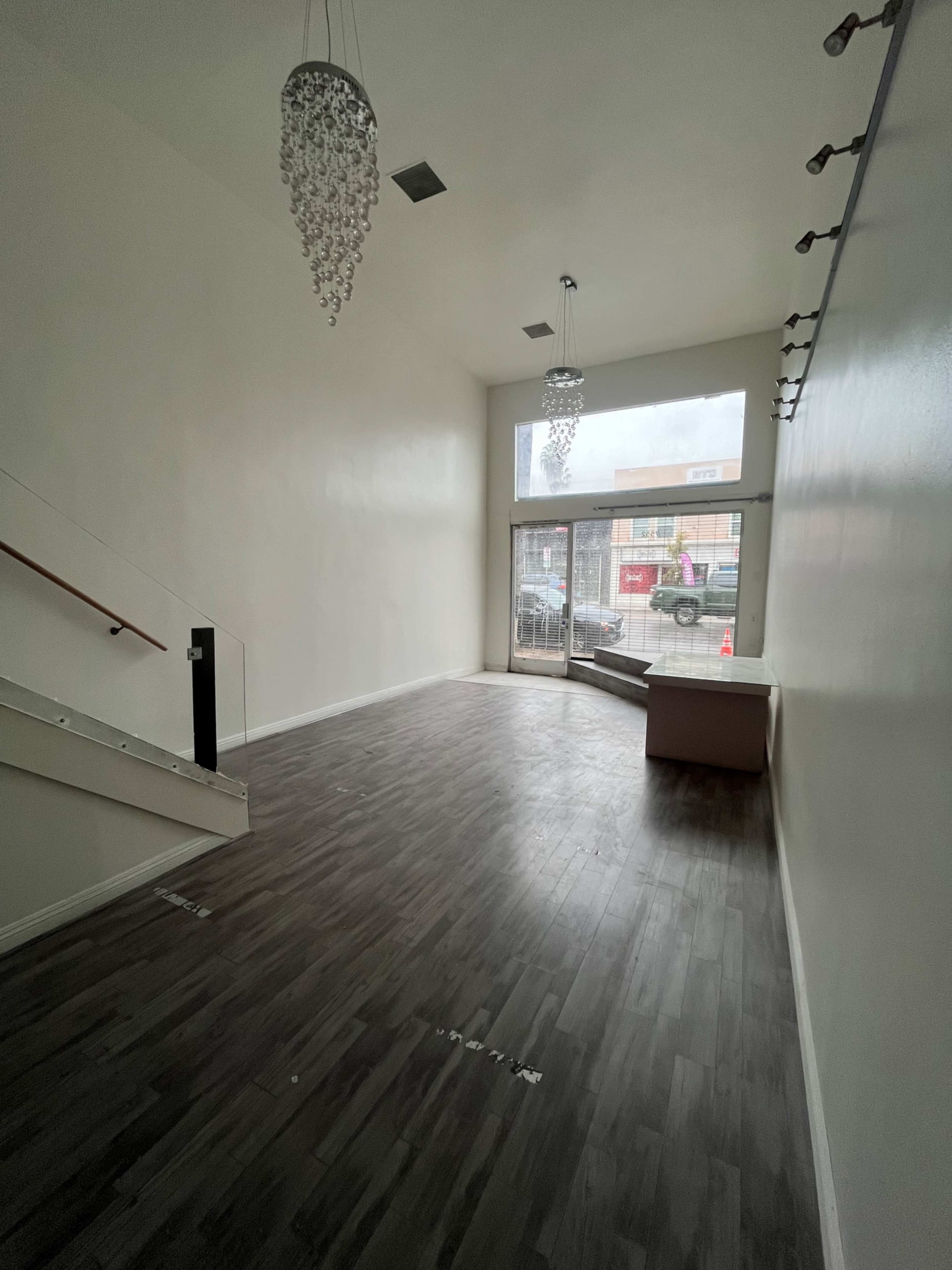 The image shows a spacious, empty retail space with large windows, modern lighting fixtures, and a wooden floor.