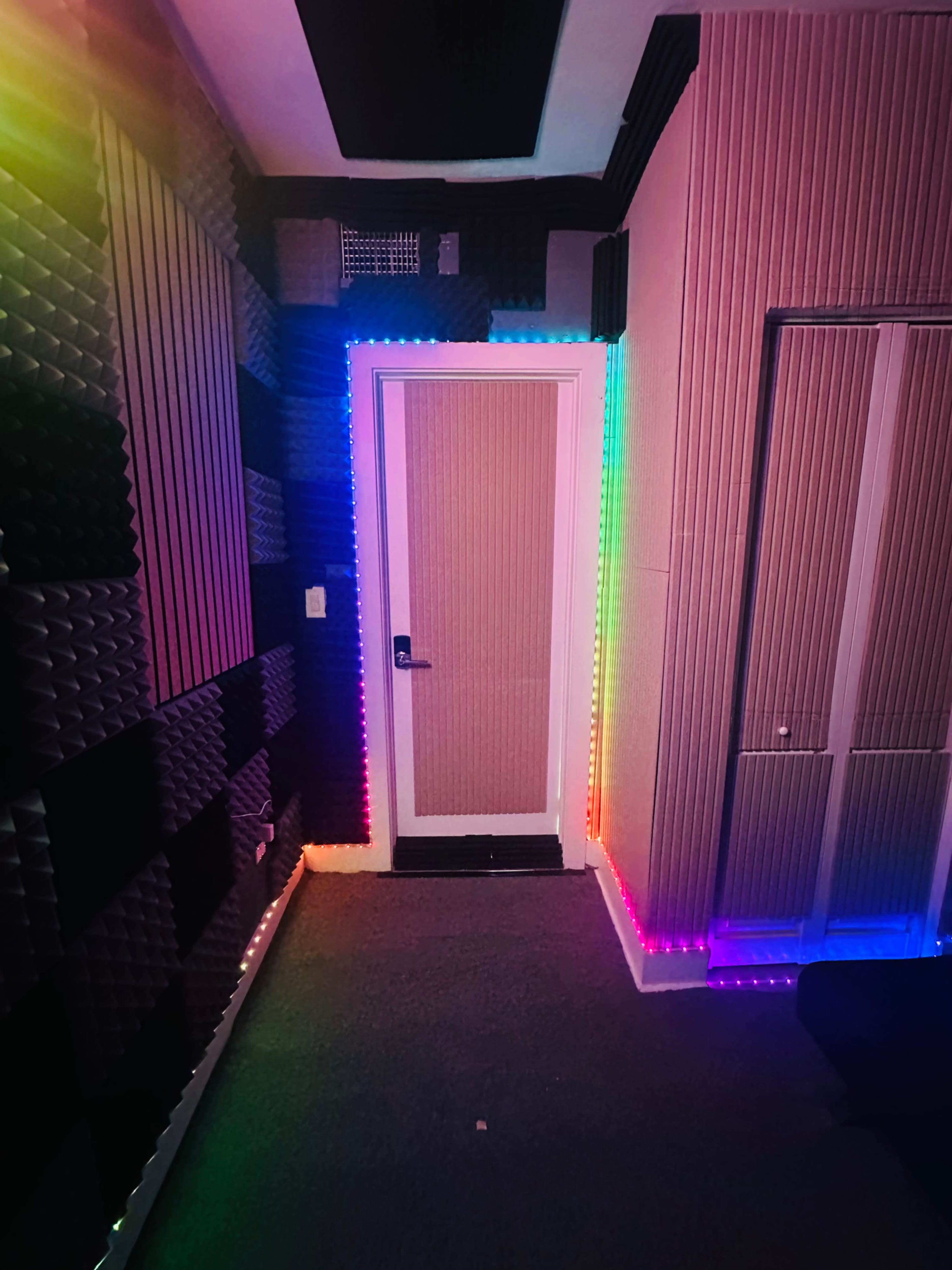 The image shows a small room with soundproofing panels on the walls and colorful LED lights outlining a doorframe.