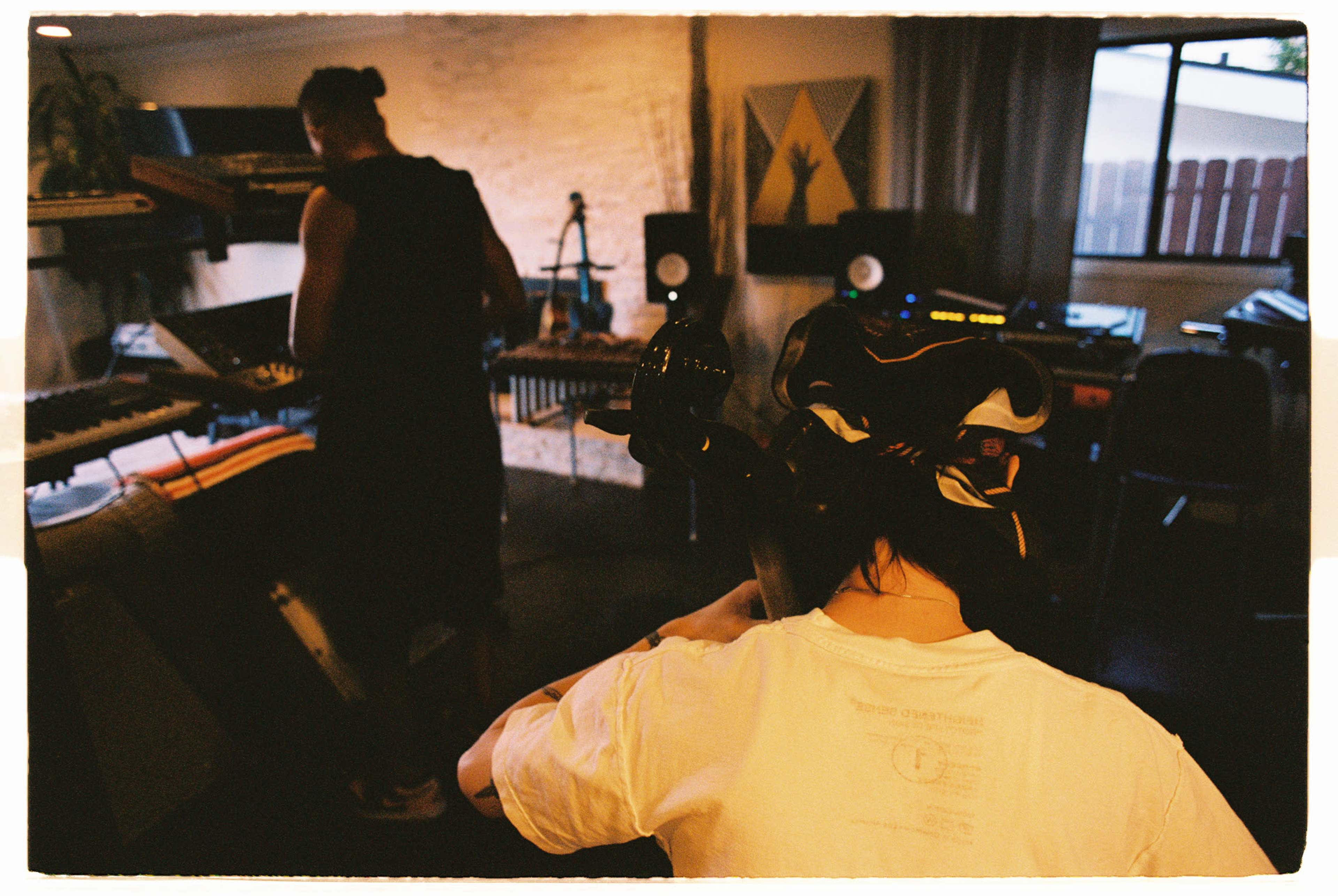 A person with a camera on their head sits in the foreground while another individual stands in the background, working with musical equipment in a studio environment.
