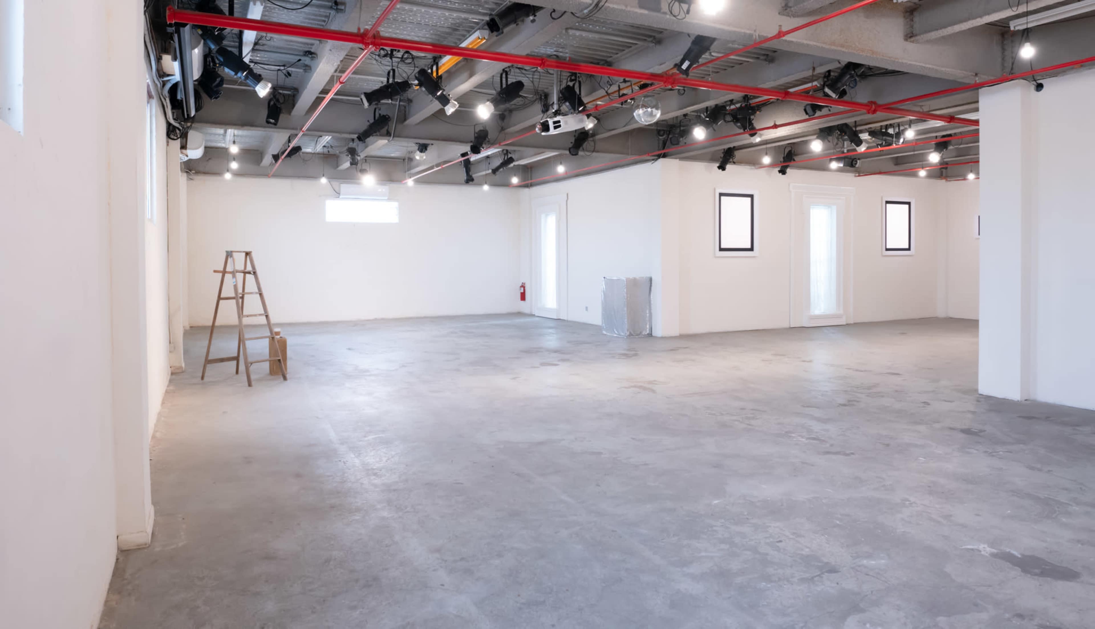 A spacious, empty room with concrete flooring, overhead lights, a ladder, and a few artworks on the walls.
