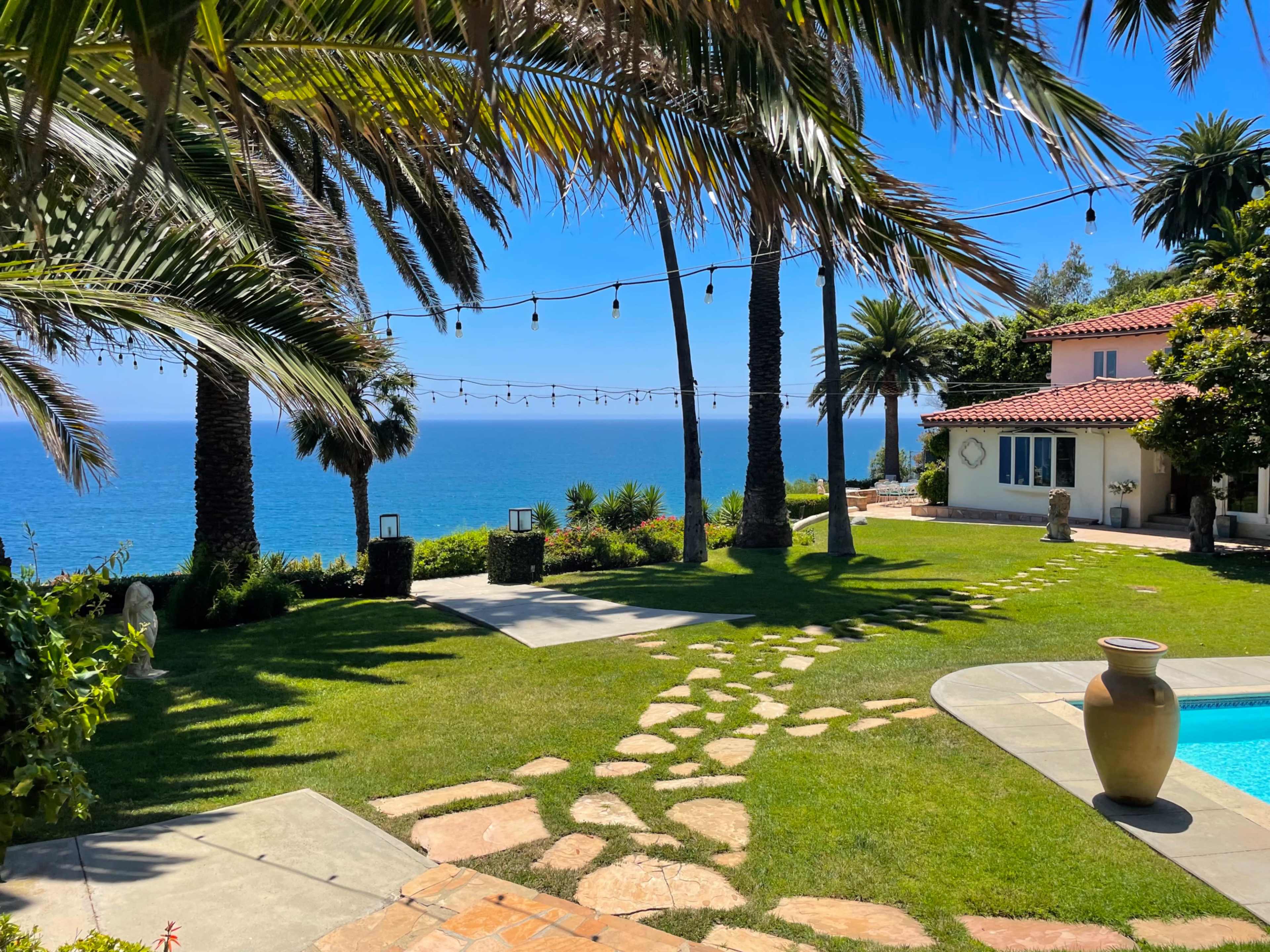 A landscaped yard features a stone pathway leading to a pool, with palm trees and ocean views in the background.