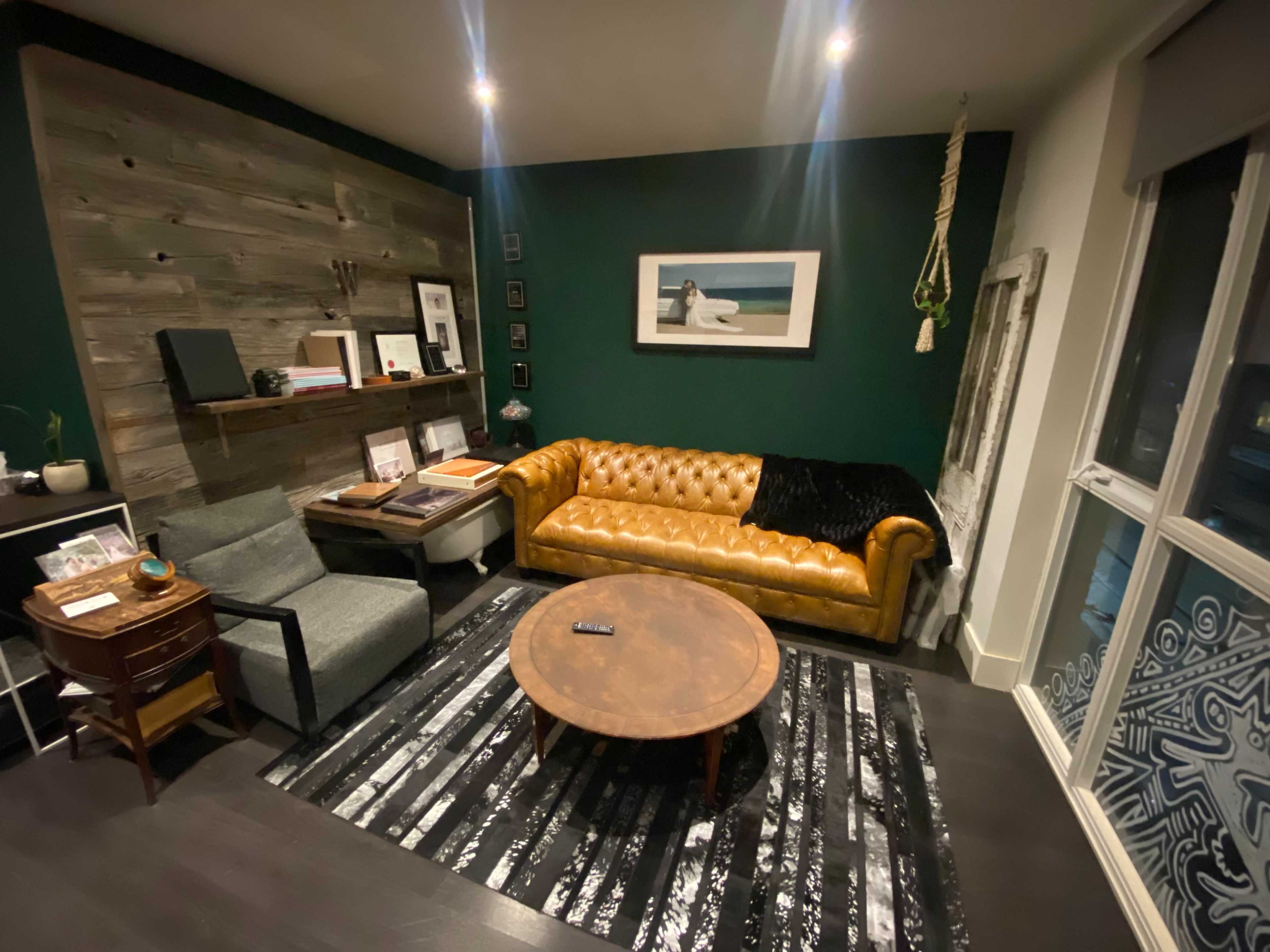 A cozy living room featuring a brown leather chesterfield sofa, a round coffee table, and a gray armchair, with a dark green accent wall and wooden paneling.