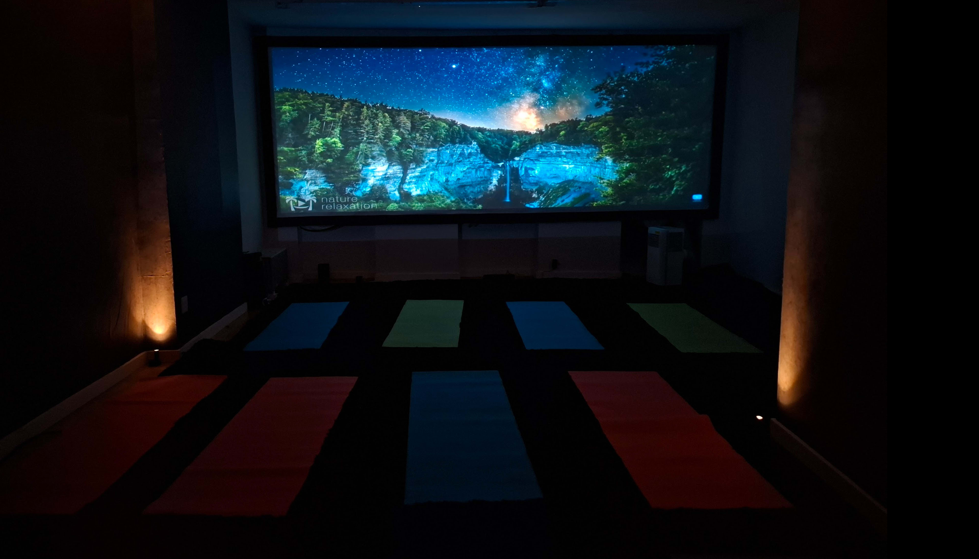 The image shows a dimly lit yoga studio with colorful mats arranged on the floor and a large screen displaying a starry night scene in the background.