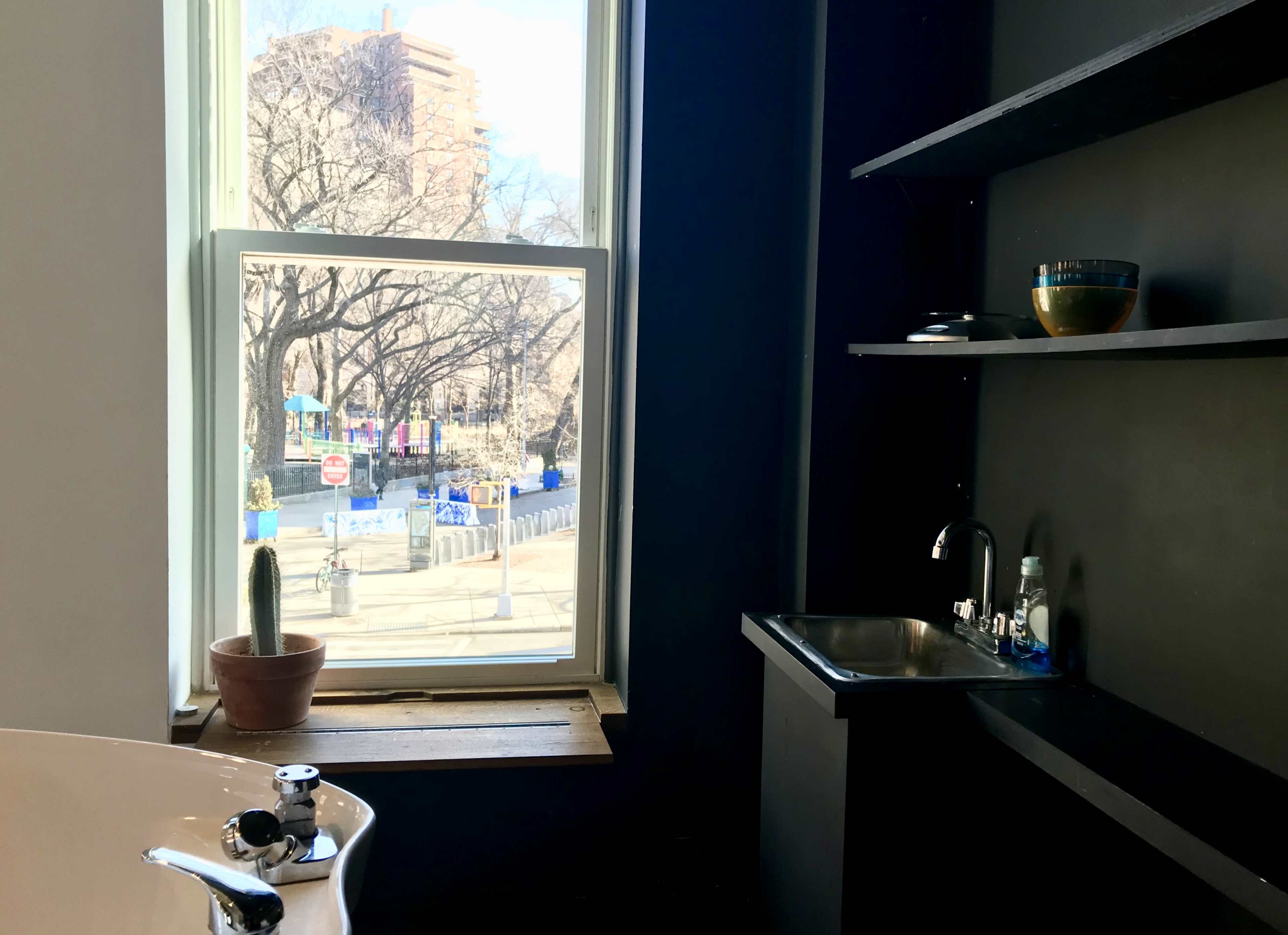 A modern bathroom with a bathtub near a window overlooking a city street, featuring a small sink and shelves.