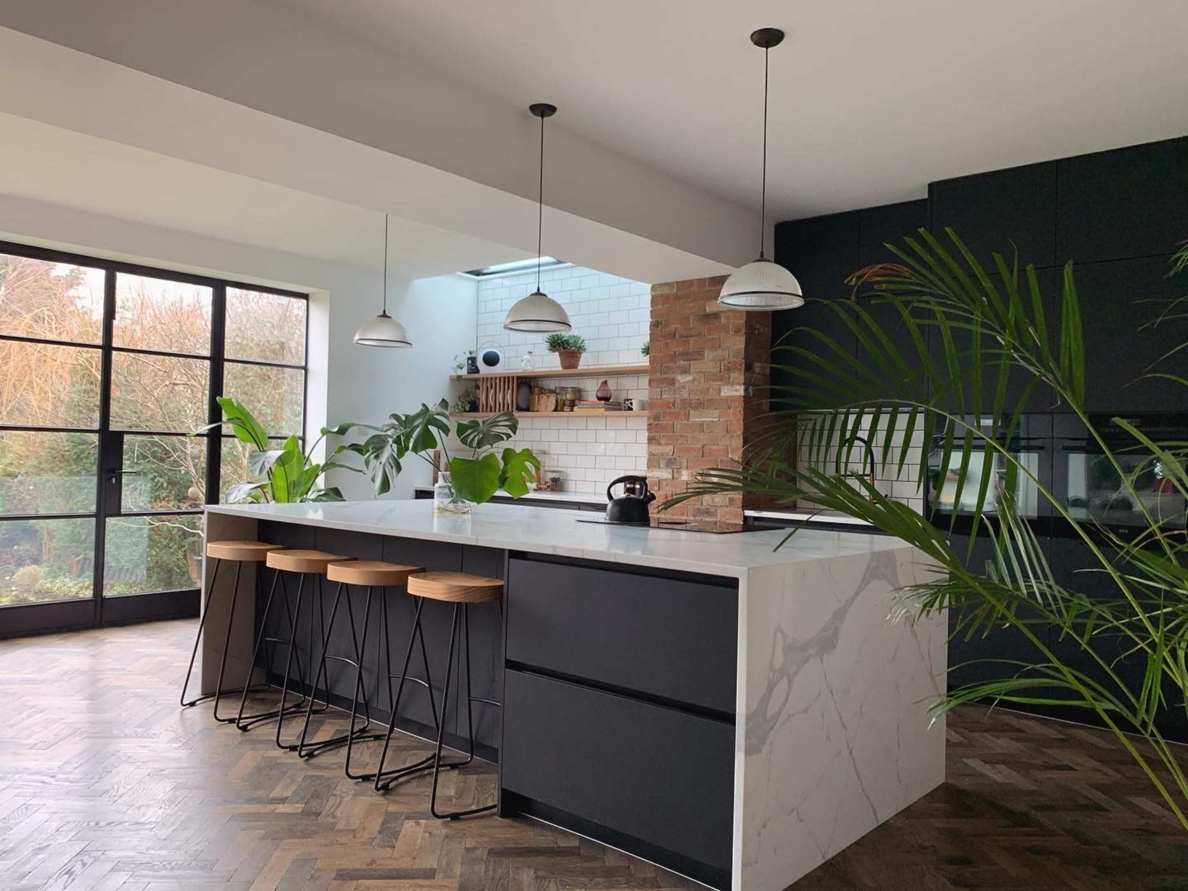 A modern kitchen with a large island, bar stools, and plants, featuring a mix of dark cabinetry and a light marble countertop.