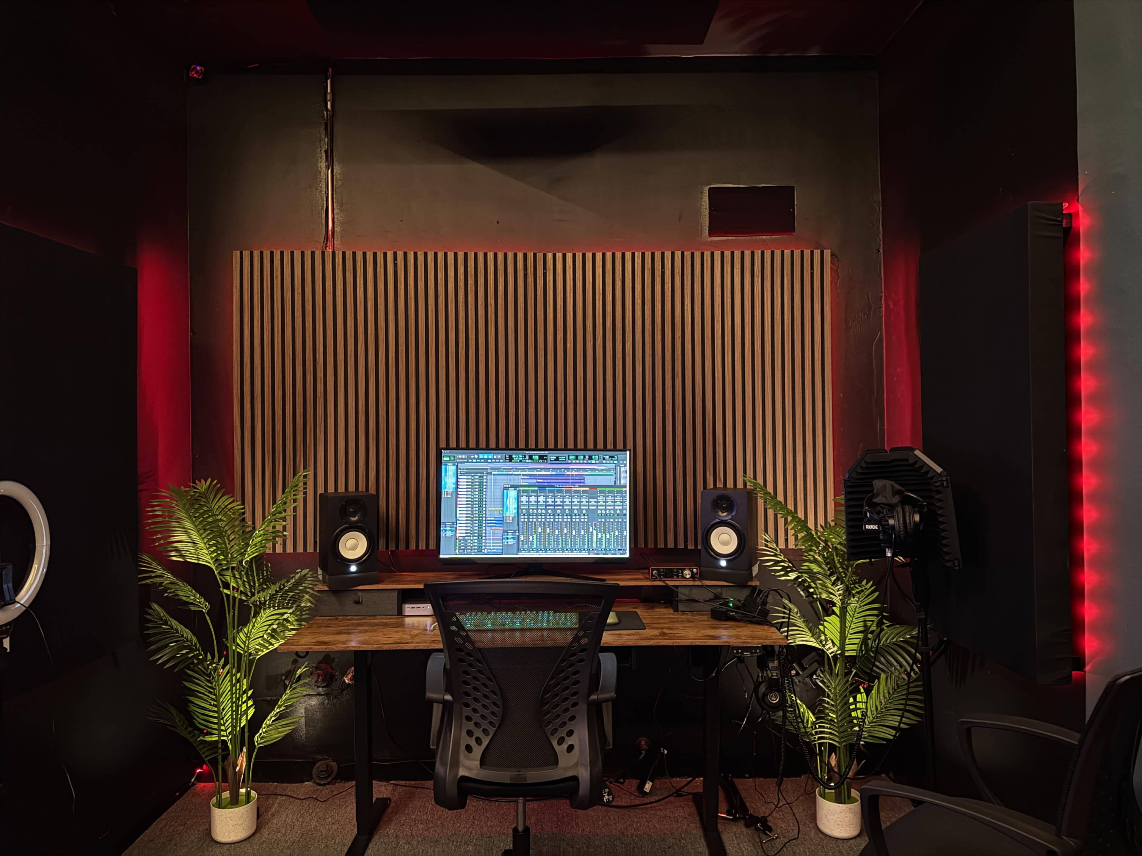 The image shows a modern audio production studio with a computer workstation, two studio monitors, and decorative plants on either side of the desk.