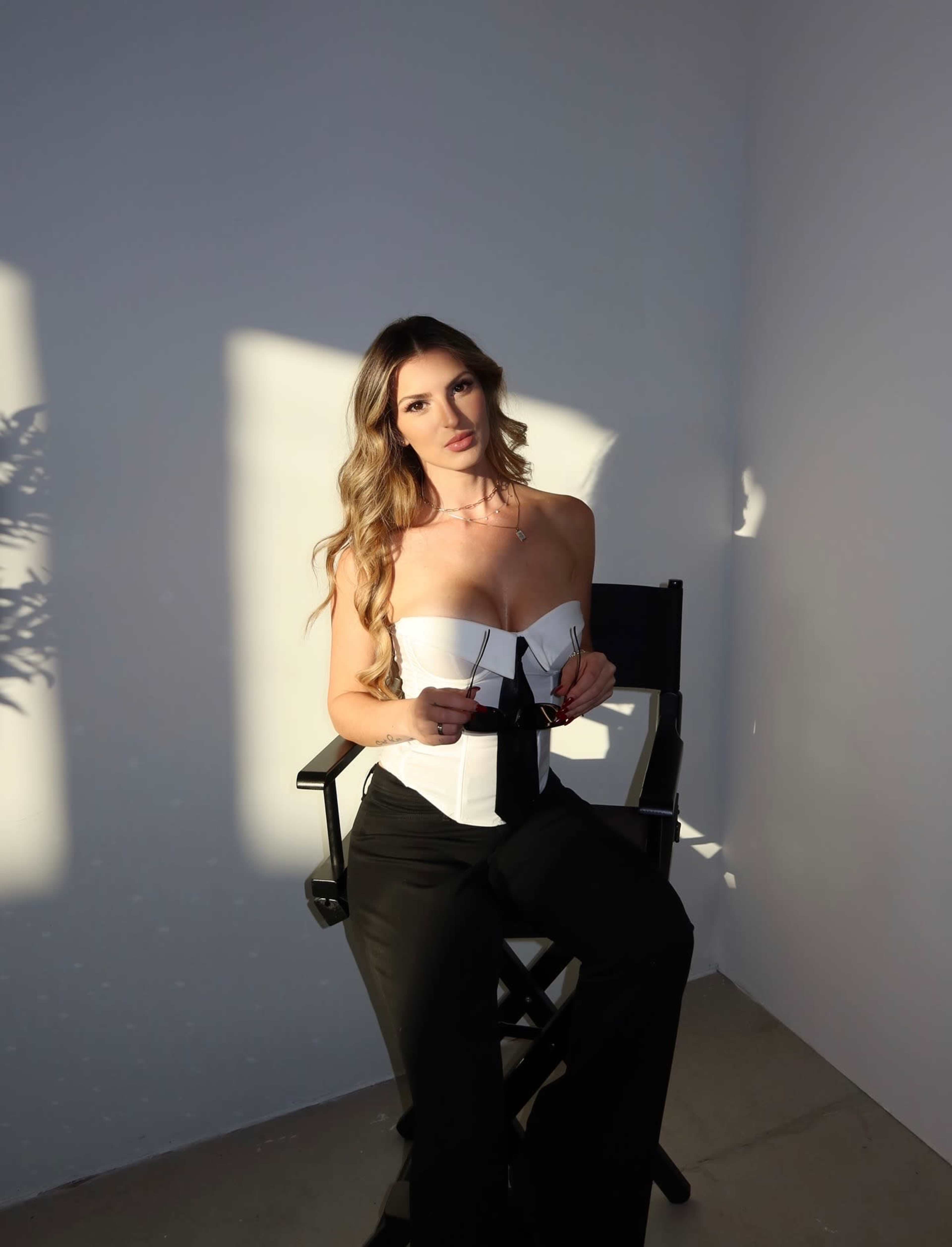A woman sits on a director's chair, wearing a strapless top and holding a pair of sunglasses, with soft light casting shadows on the wall behind her.