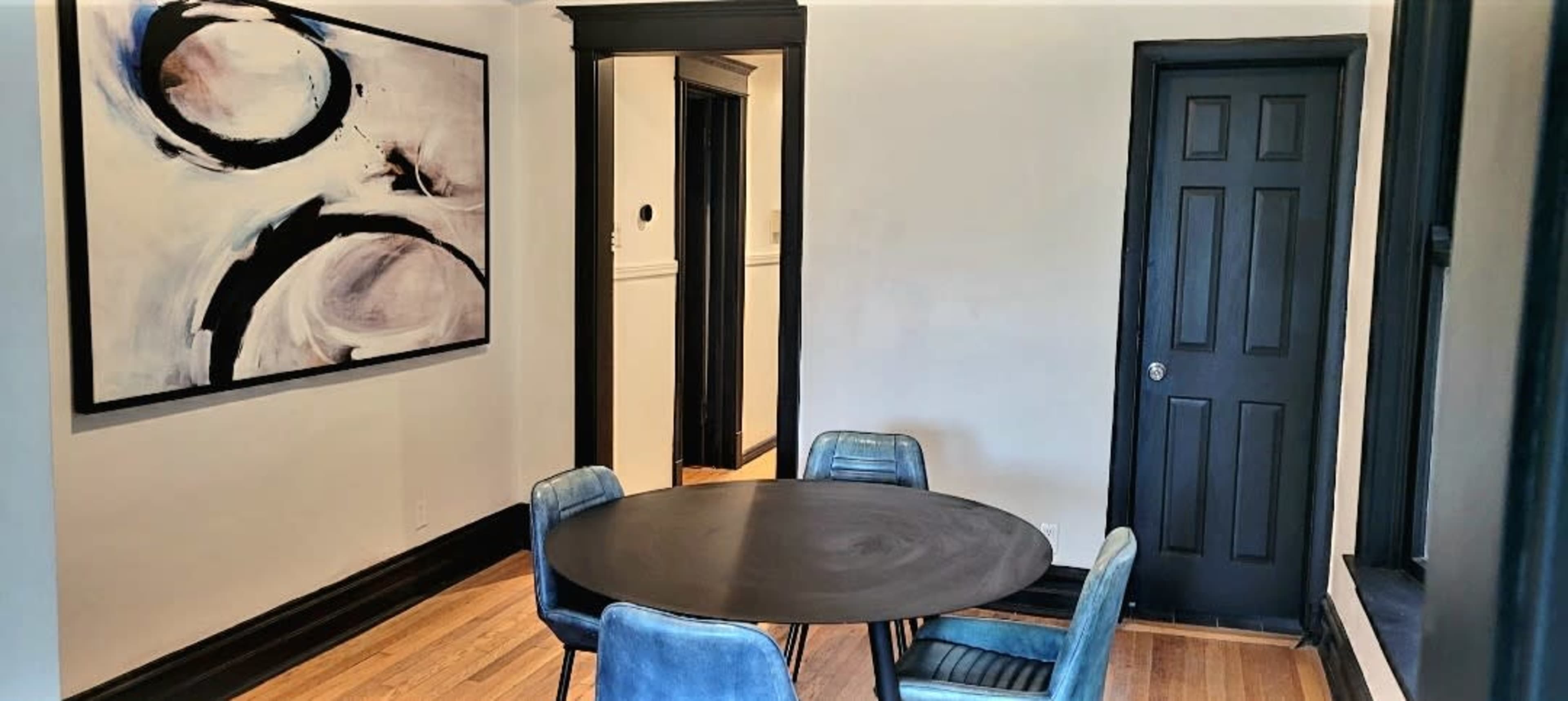 A round black table is set in a brightly lit room with four blue chairs and an abstract painting on the wall.