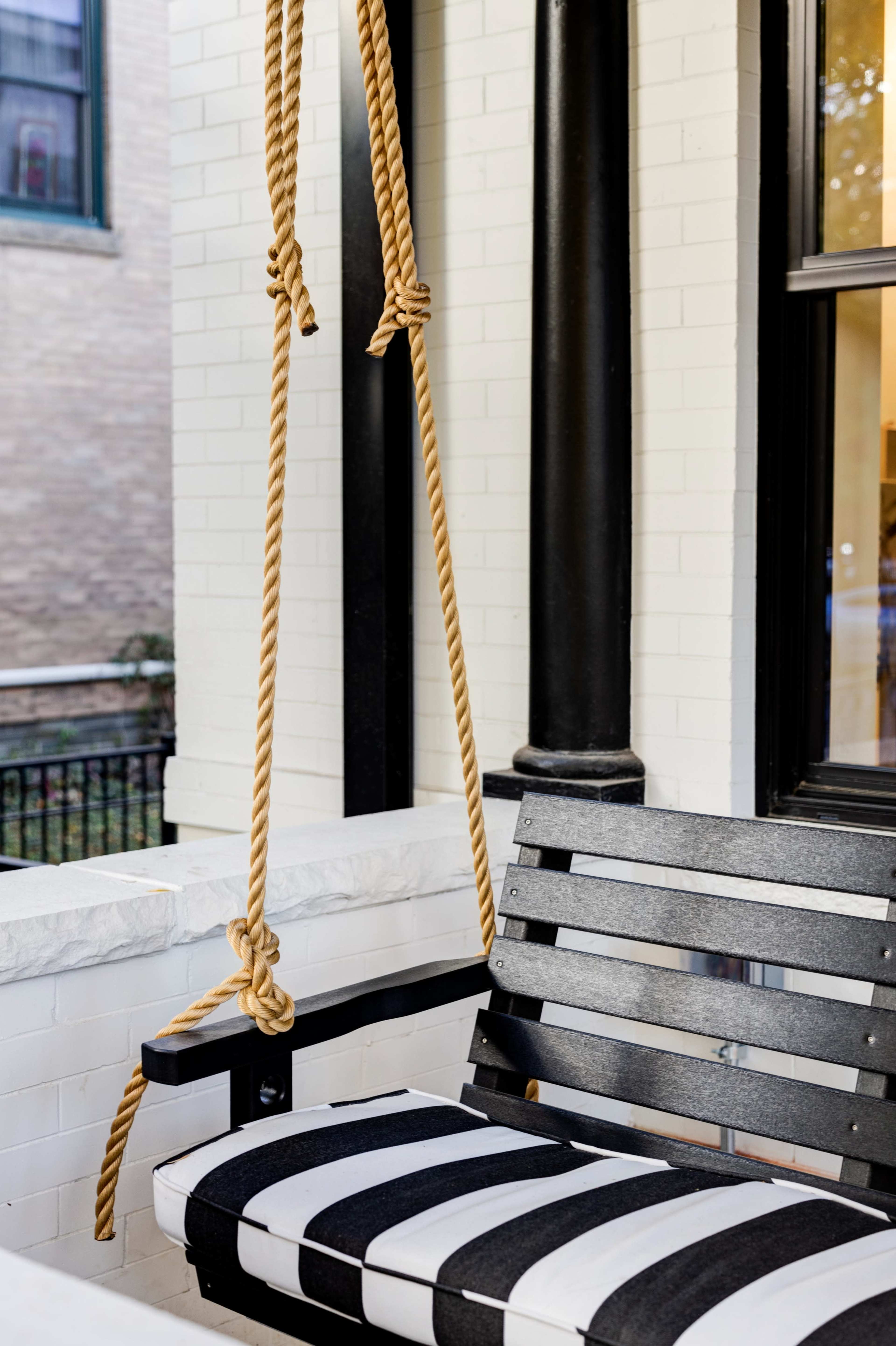 A black and white striped swing seat hangs from ropes on a porch with white brick walls and a black column.