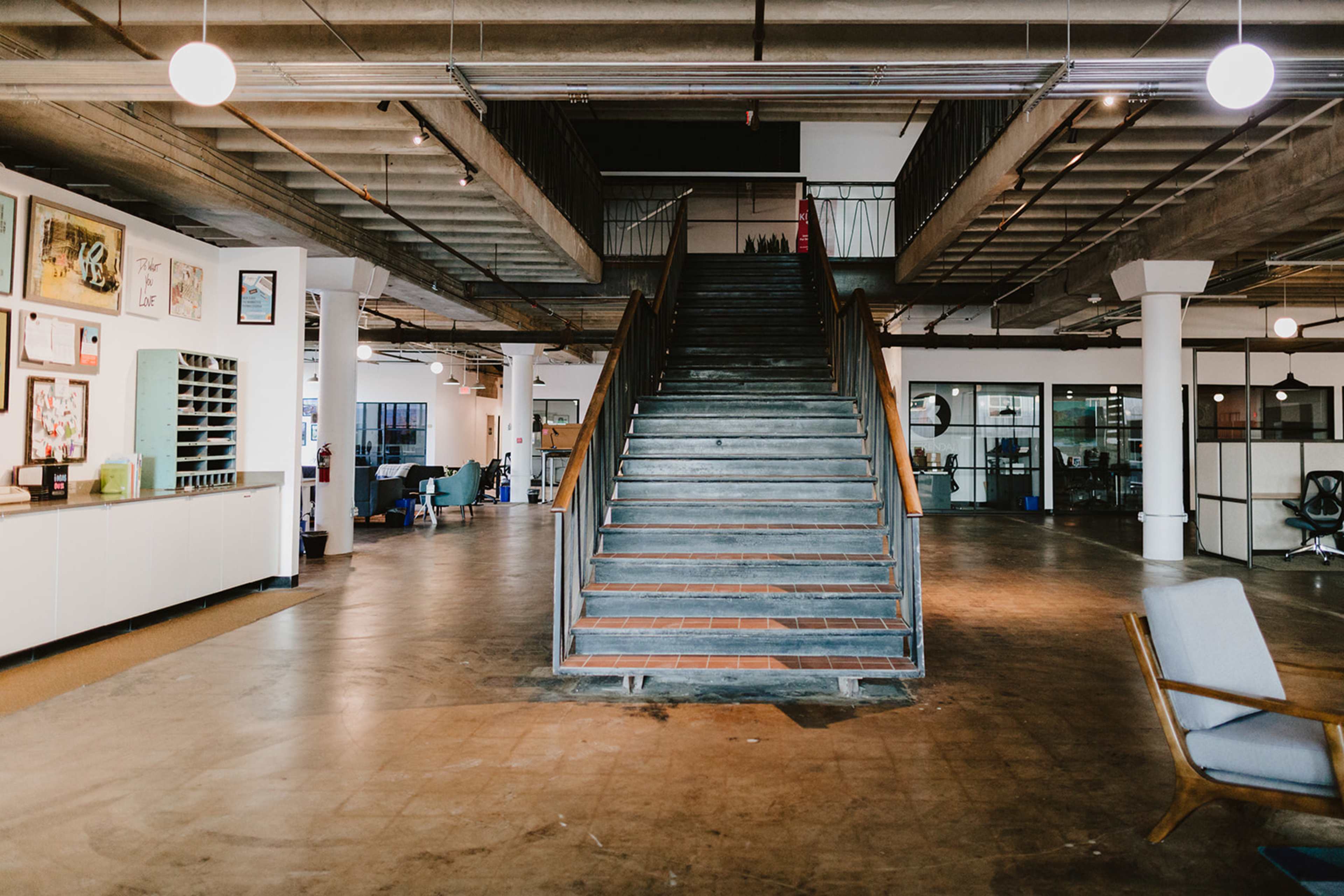 The image shows a spacious, modern office interior featuring a prominent staircase, artwork on the walls, and various seating areas.