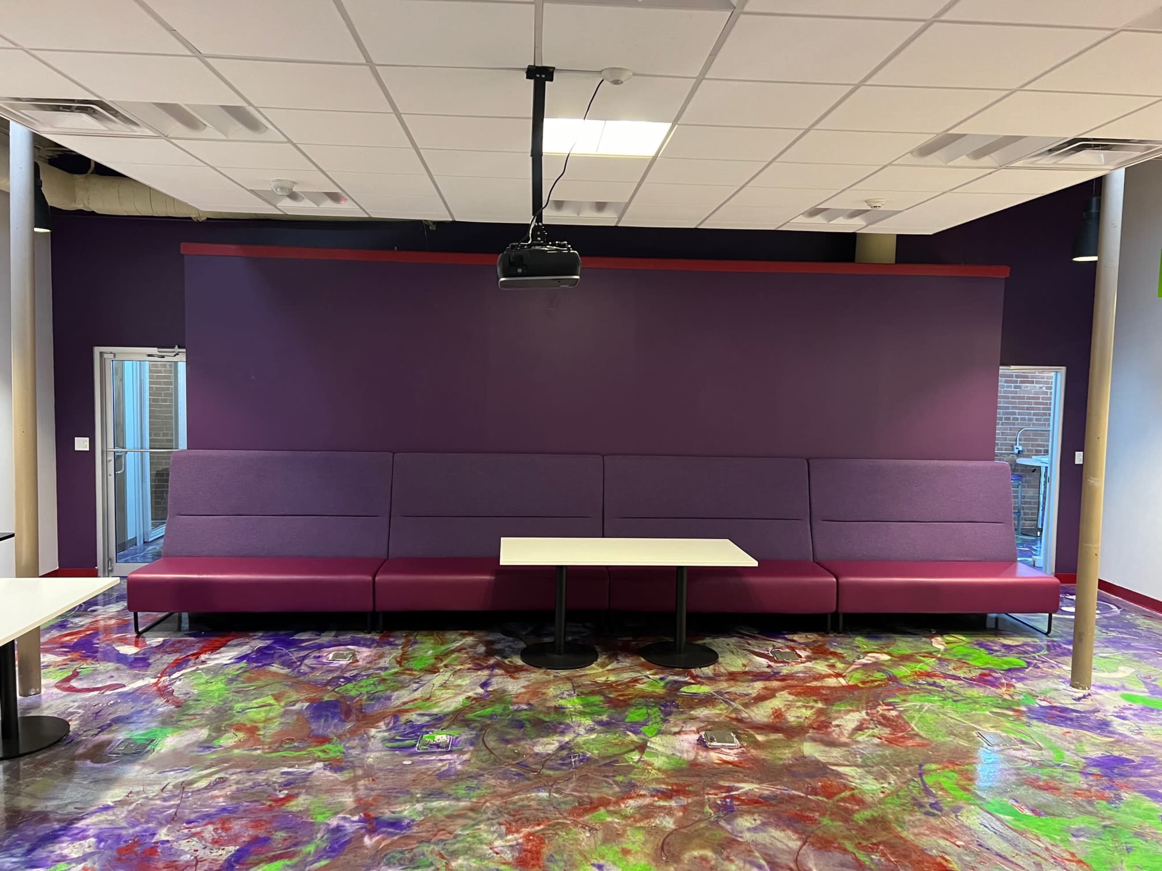 A seating area with a purple wall, purple cushioned benches, and a white table on a colorful floor.