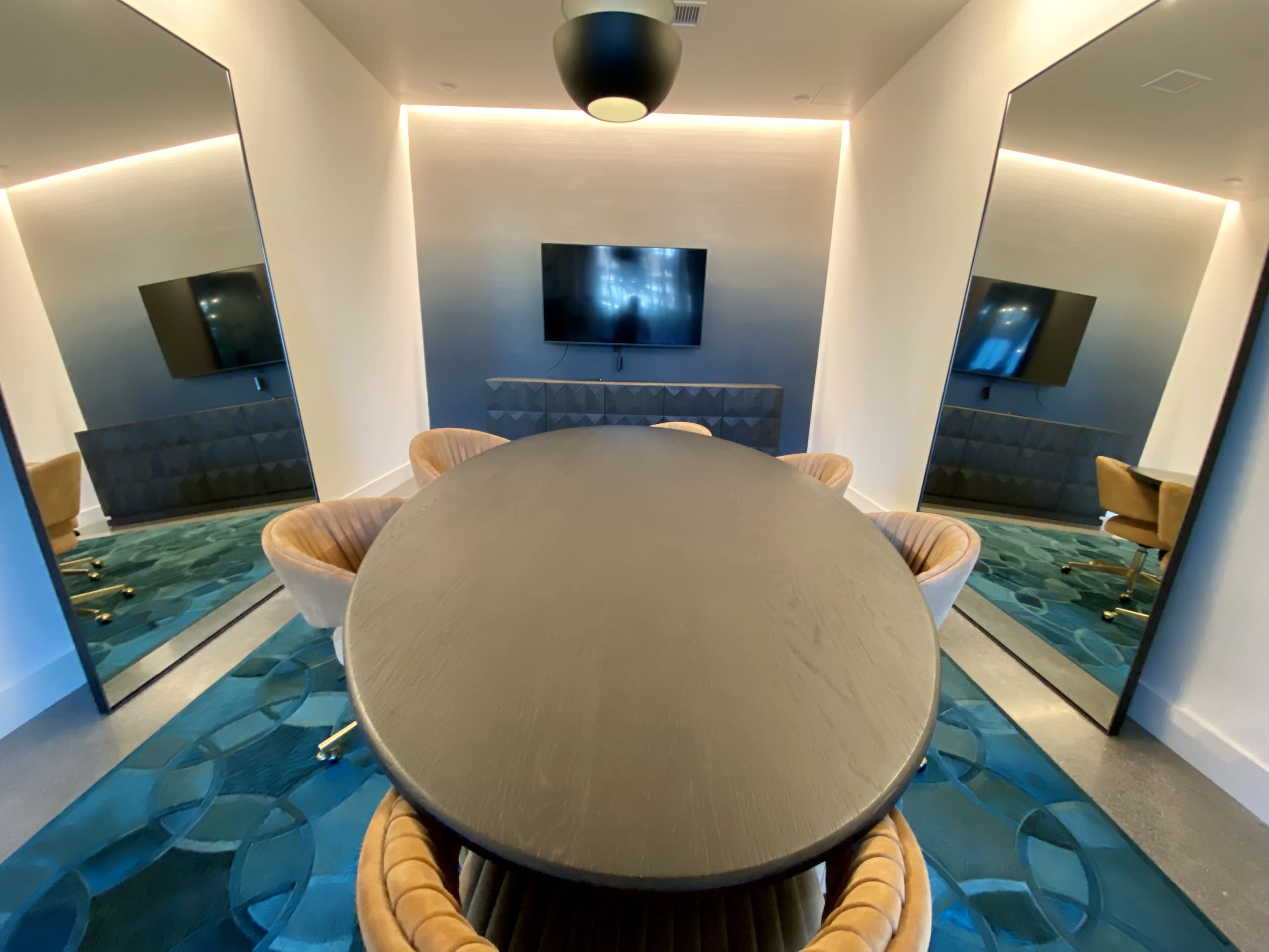A modern meeting room features a round table surrounded by four chairs, a television mounted on a wall, and two large mirrors reflecting the space.