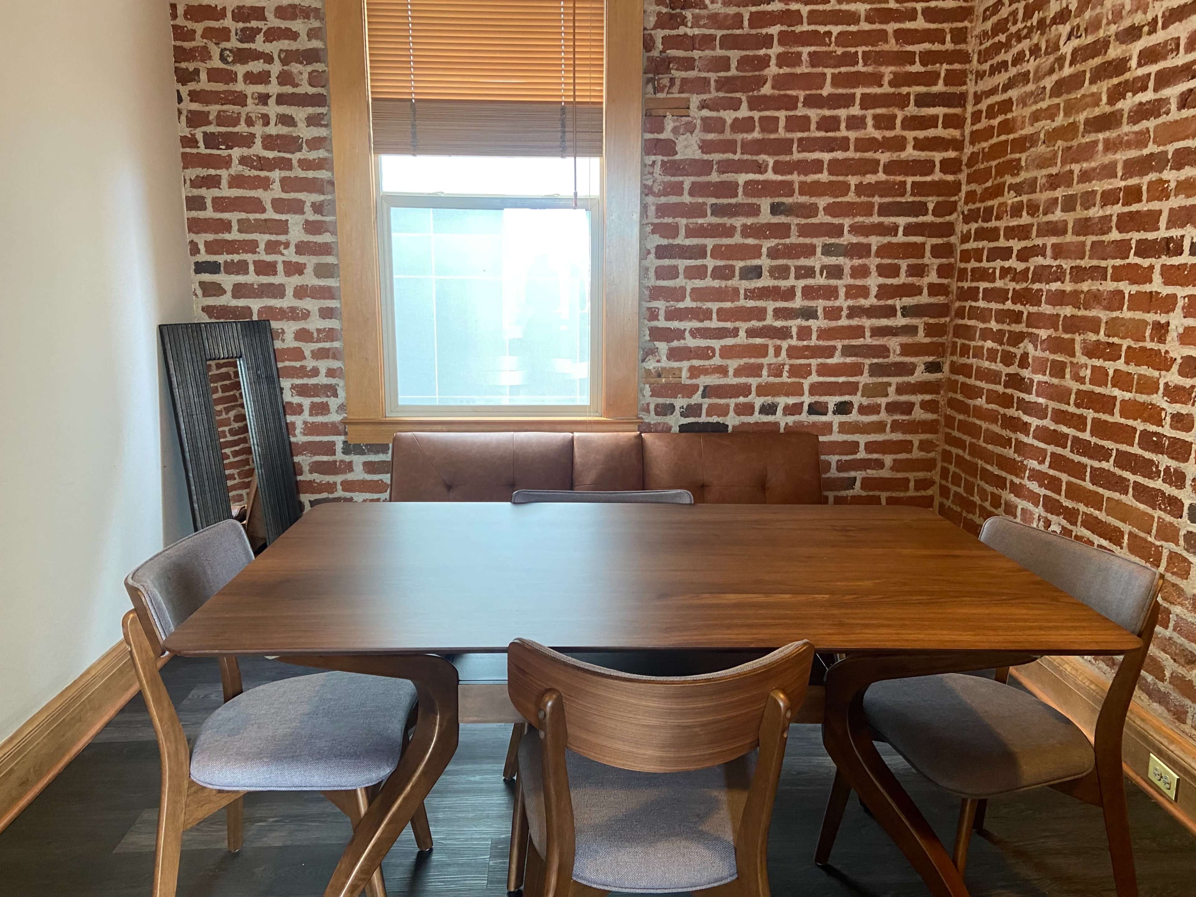 A wooden dining table with four chairs is positioned in a room featuring exposed brick walls and a window with wooden blinds.