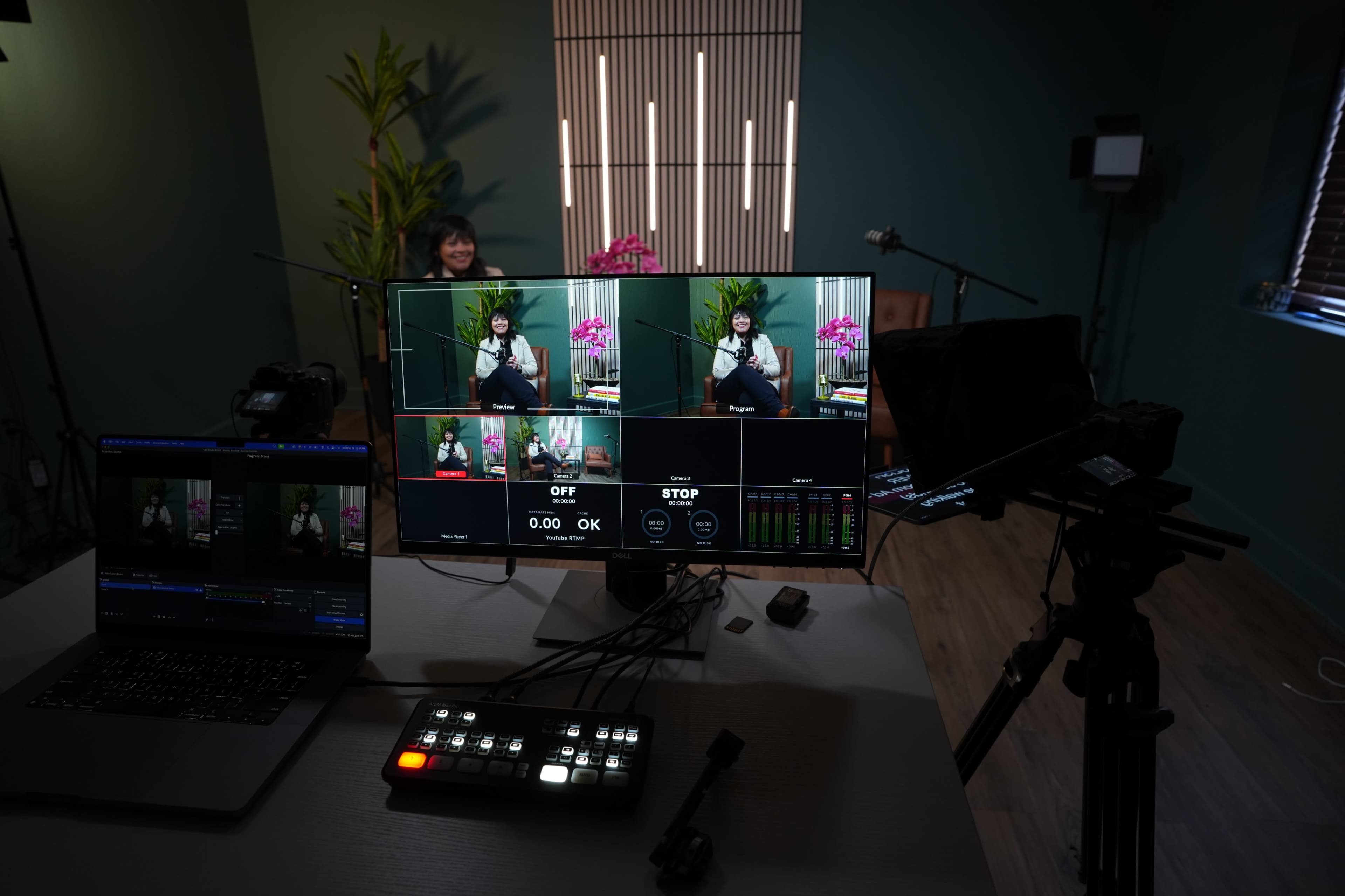 The image shows a media setup with multiple screens displaying different camera angles of a person seated in a studio, surrounded by plants and lighting equipment.