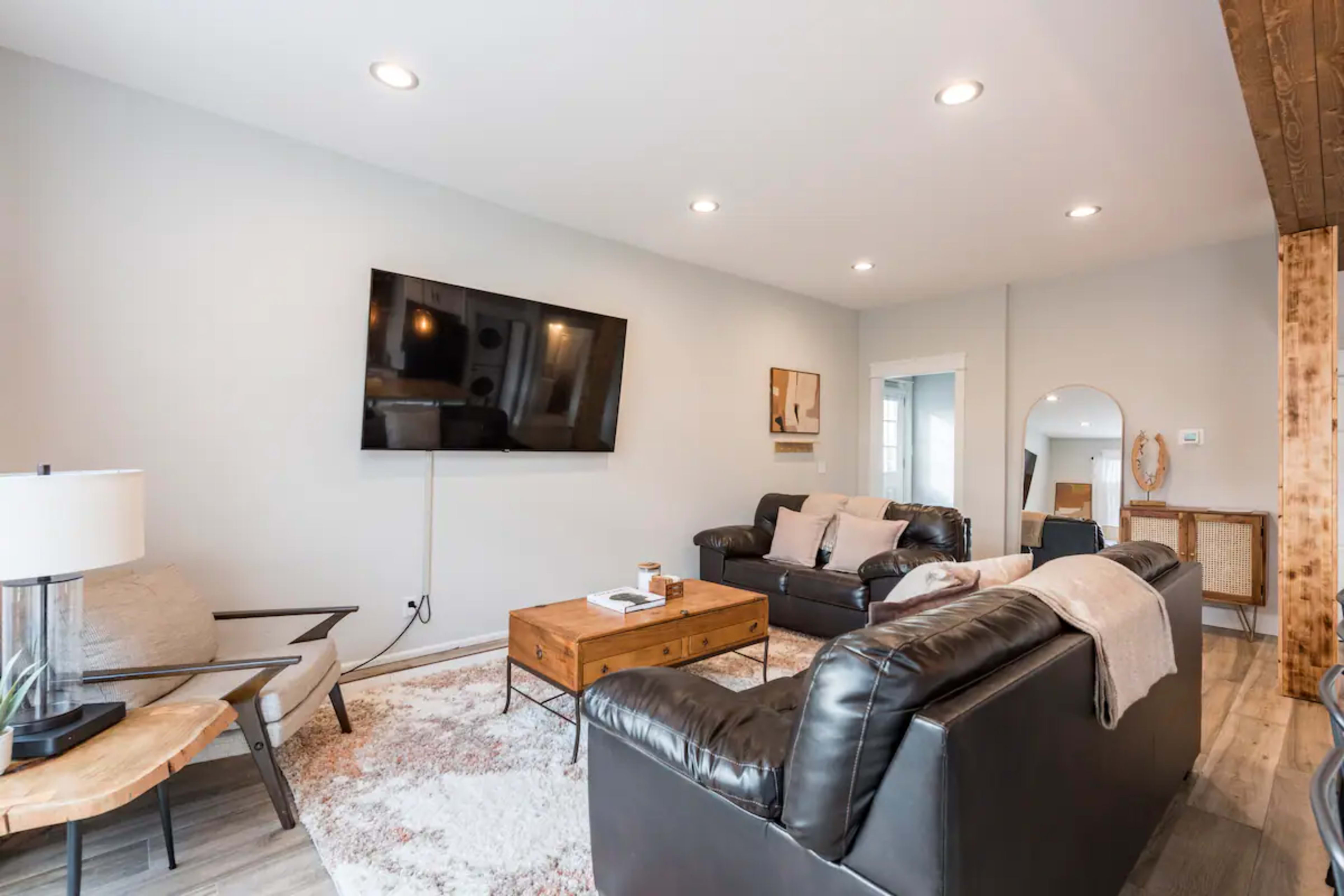 The image shows a modern living room featuring a black leather sofa, a wooden coffee table, a wall-mounted television, and neutral-colored walls with recessed lighting.