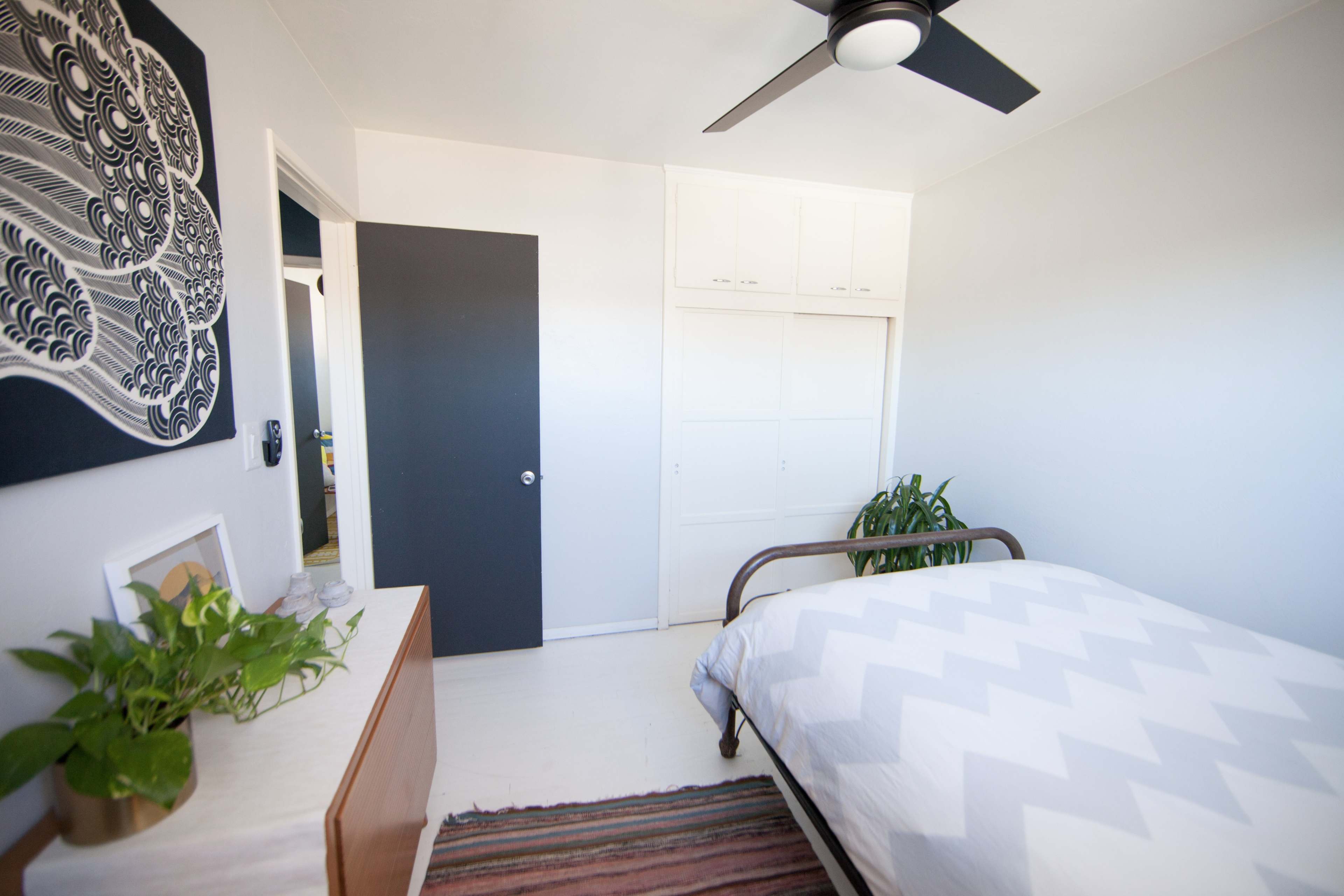 A bright bedroom features a bed with a chevron-patterned comforter, a dresser, a potted plant, and a closet door against neutral-colored walls.