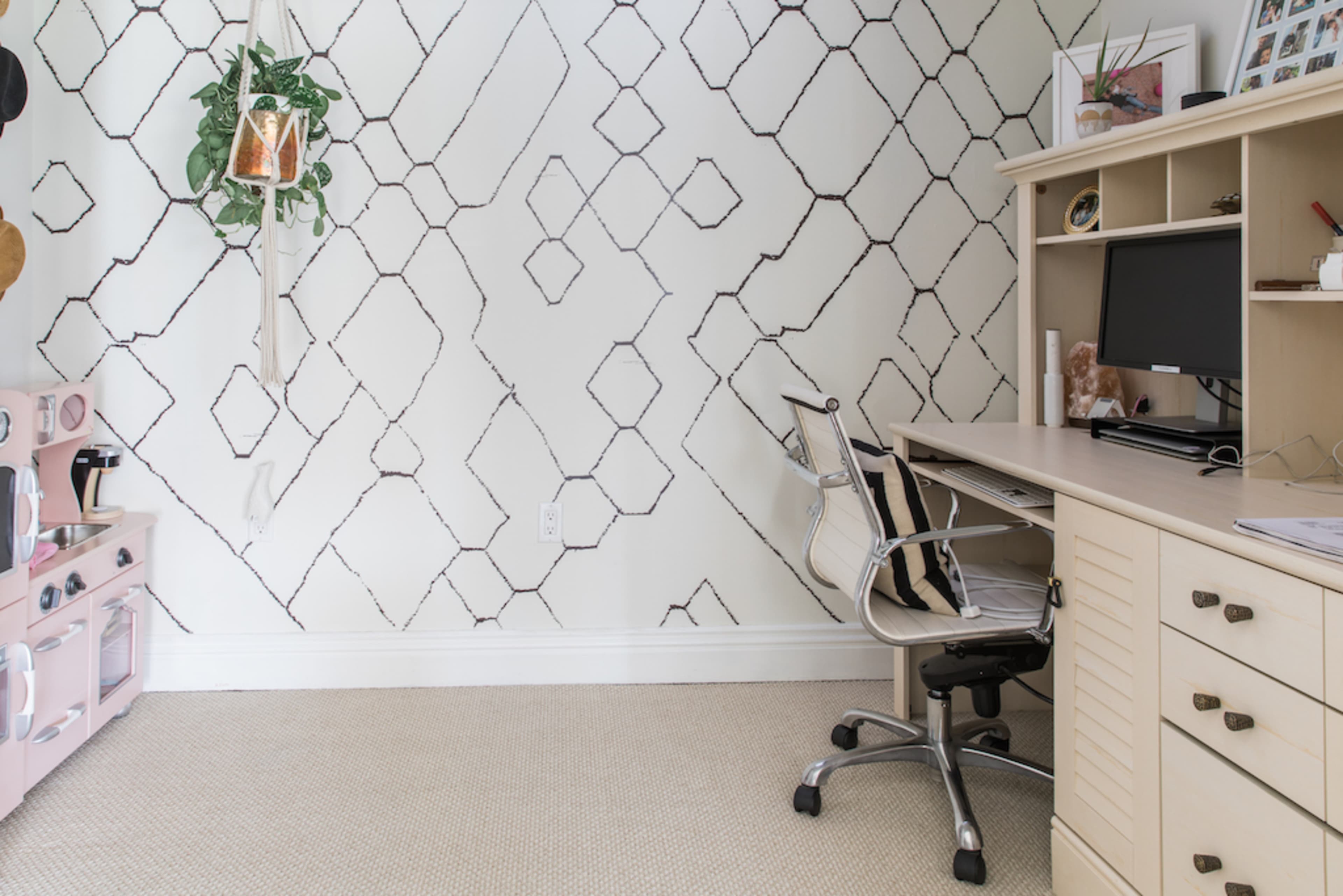 A home office features a light-colored desk with a computer chair, surrounded by a geometric patterned wall and a small hanging plant.