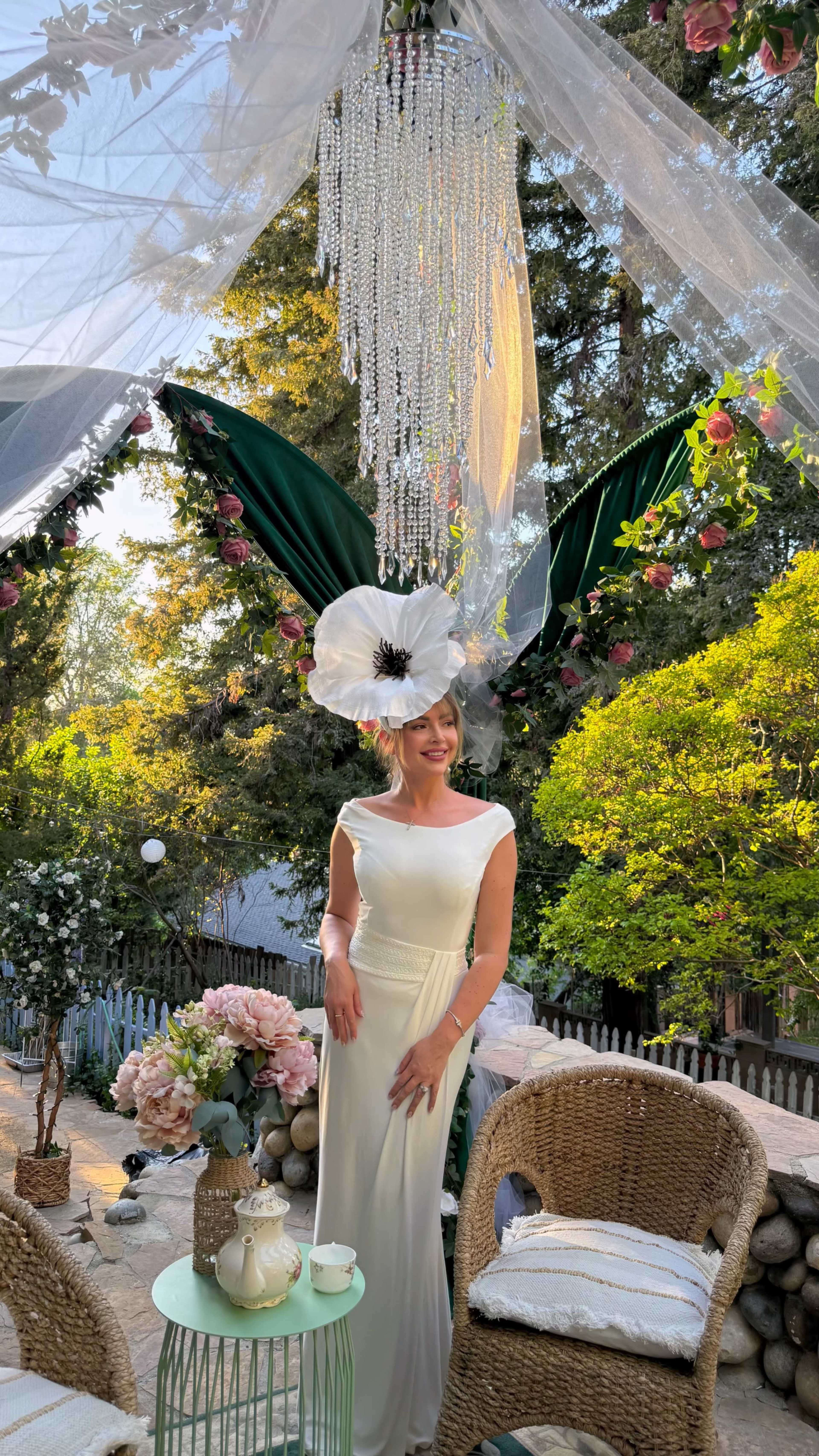A woman in a white dress stands in a garden setting, adorned with greenery and floral decorations, under a floral arrangement hanging above her.