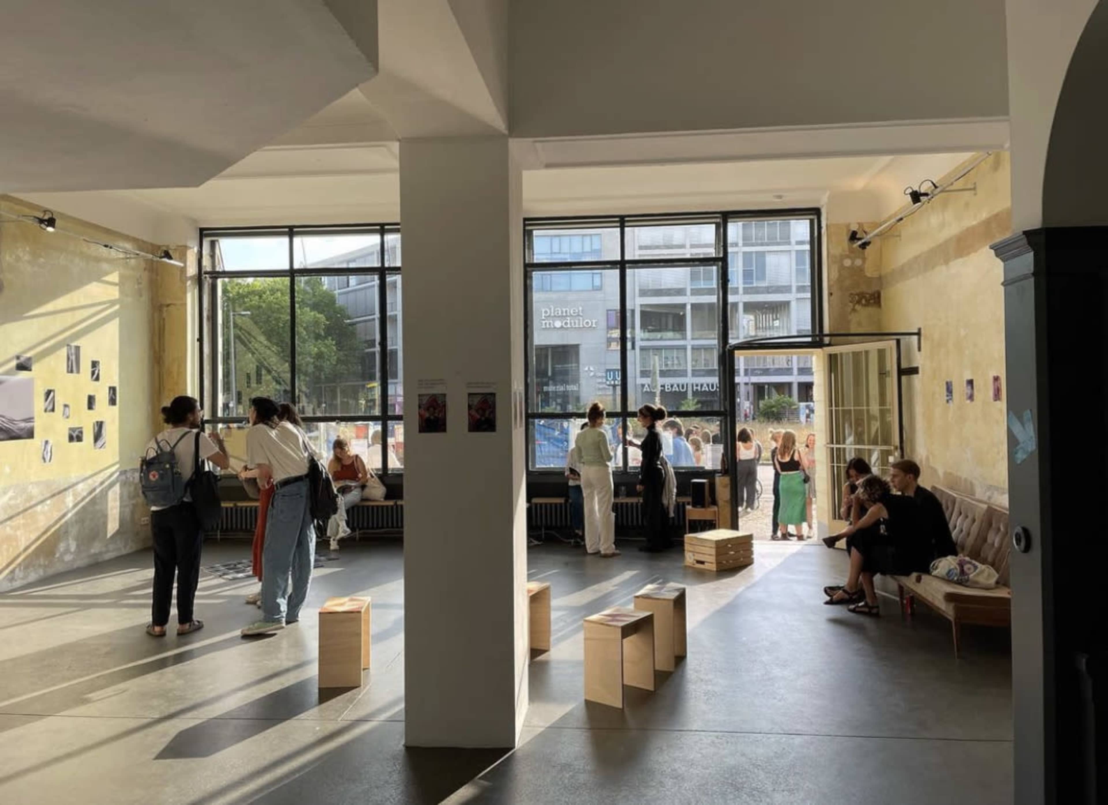 The image shows a spacious art gallery with large windows, where several people are engaged in conversation and viewing displayed artworks.