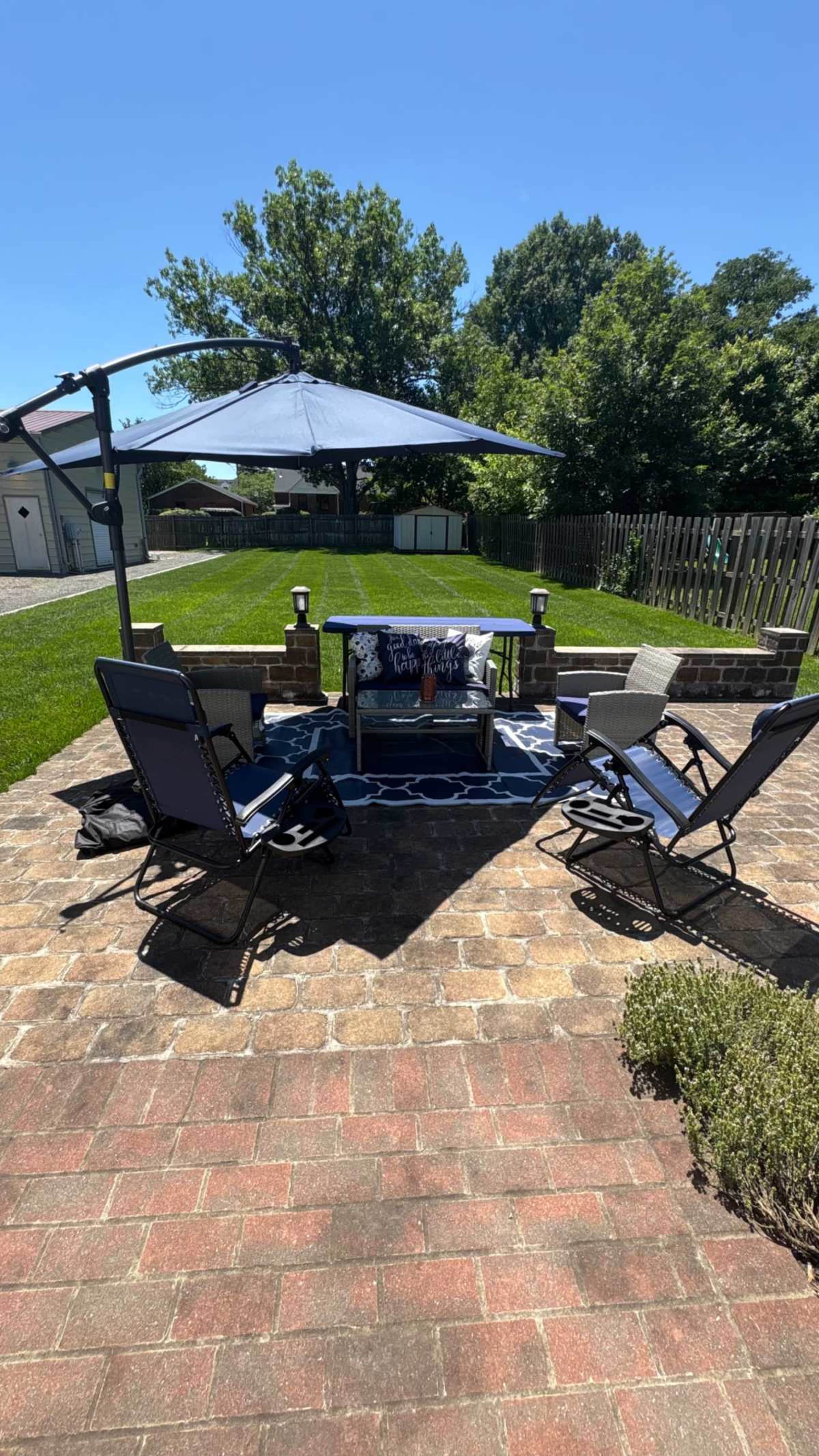 The image shows a patio area with four folding chairs arranged around a table, set under an umbrella, surrounded by a lawn and a wooden fence.