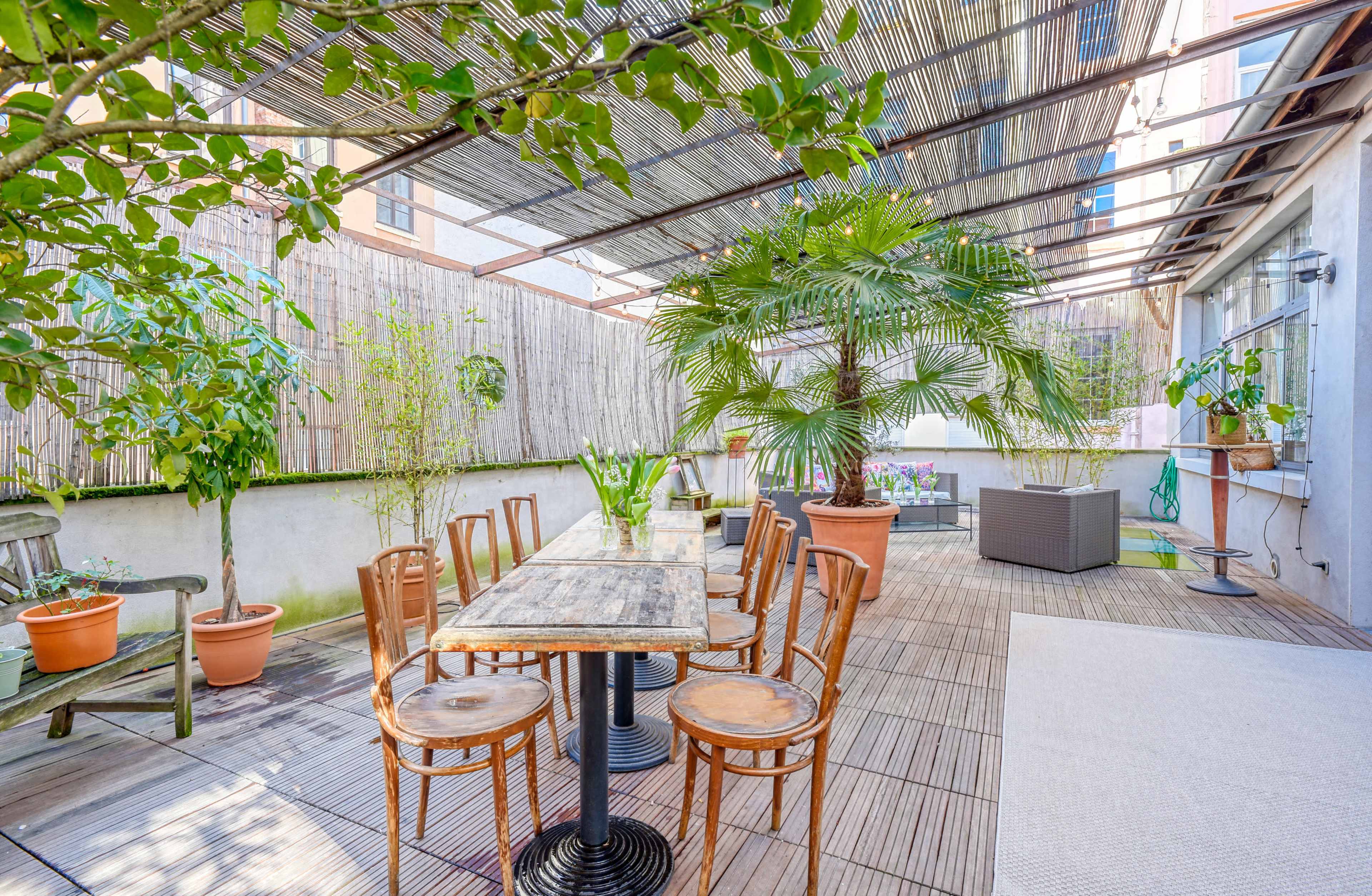 The image shows a spacious rooftop terrace covered with a wooden pergola, featuring potted plants and a wooden table surrounded by chairs.