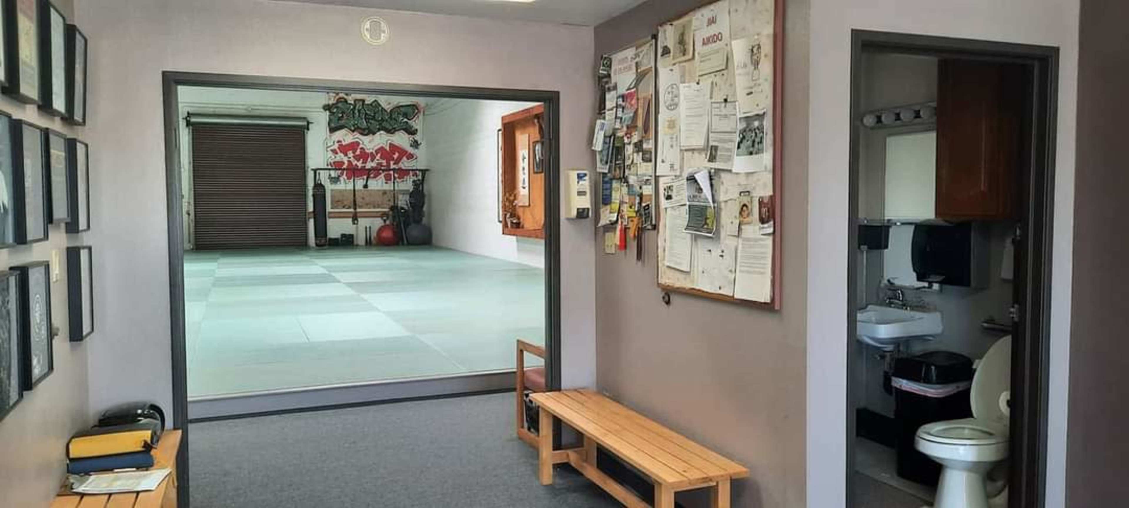 A hallway leading to a dojo, with a bulletin board on one wall and a bench in the foreground.