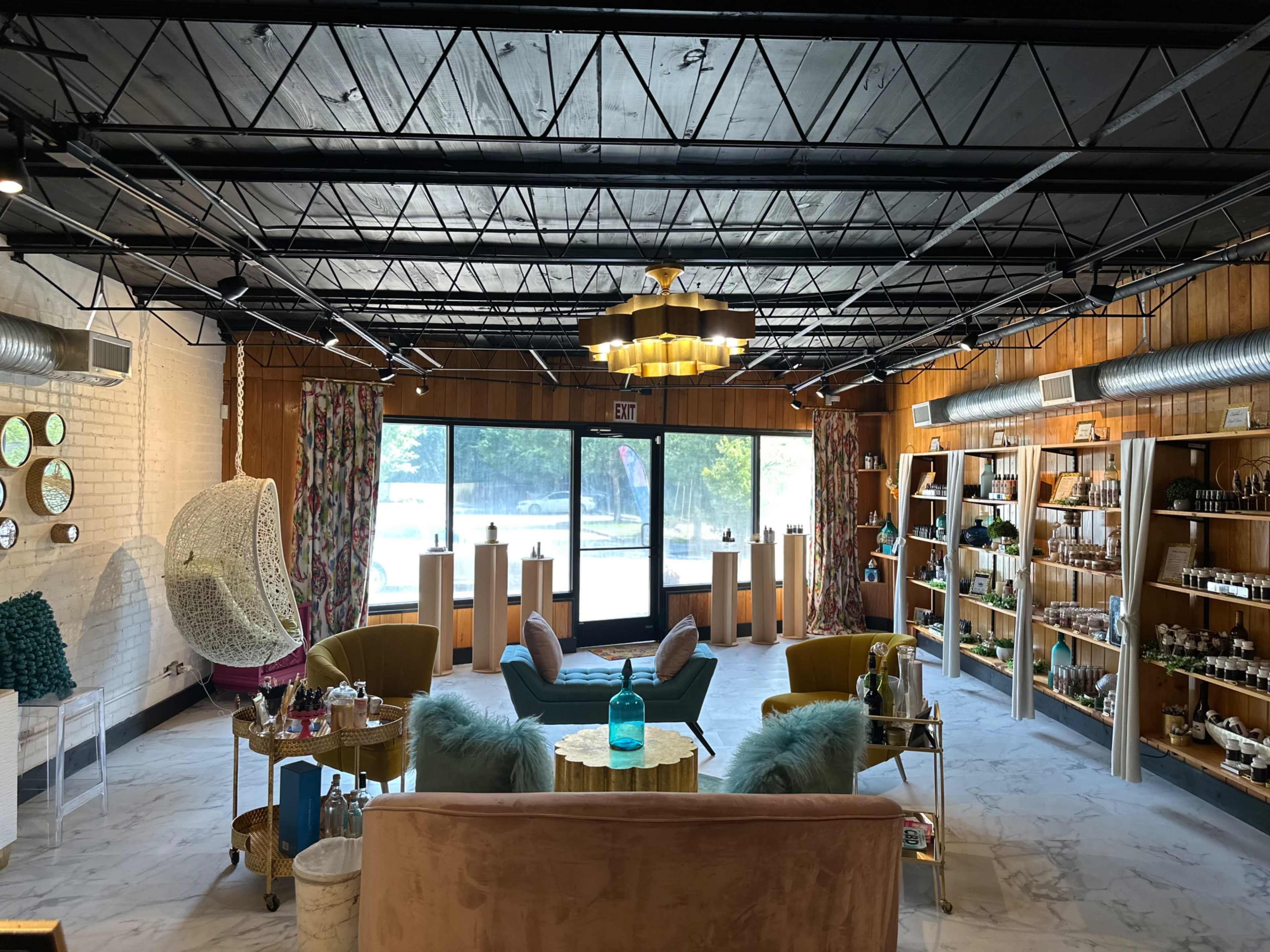 The image shows a modern interior of a beauty or wellness store with seating areas, decorative shelves filled with products, and large windows letting in natural light.