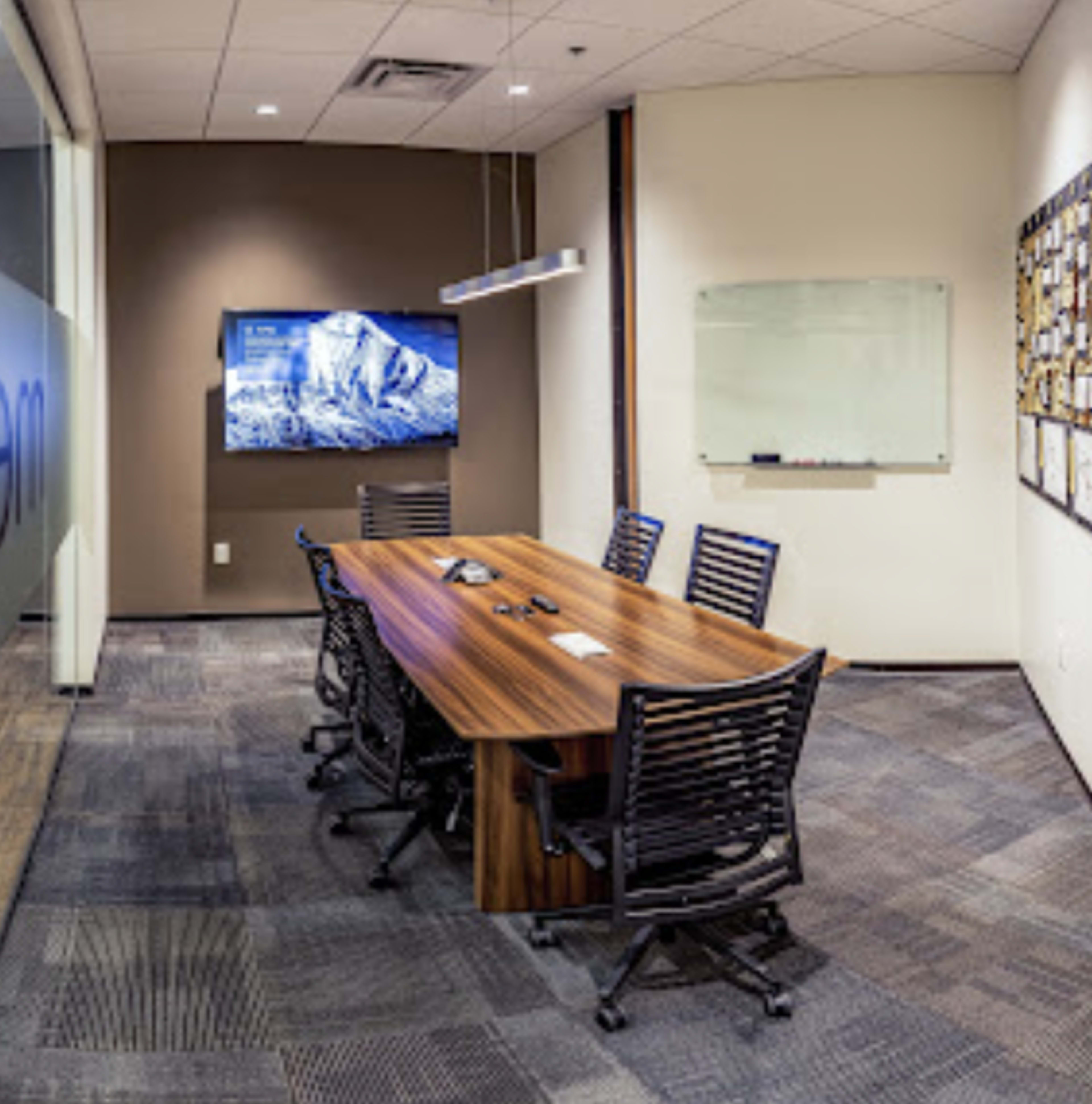 A conference room features a long table surrounded by chairs, a television display on the wall, and a whiteboard.